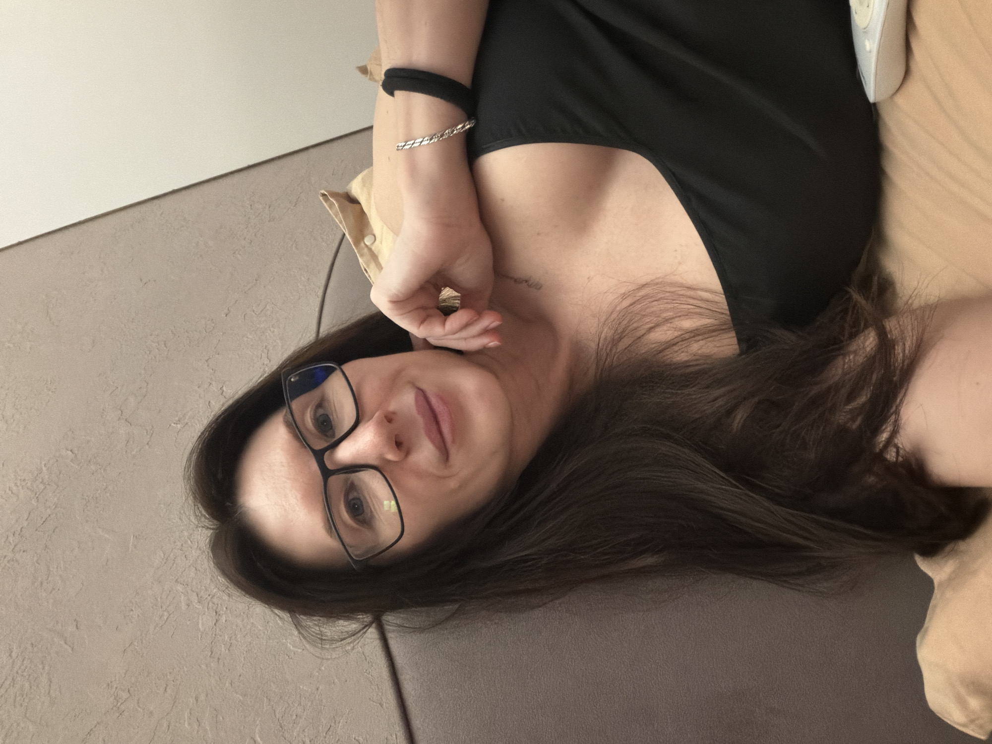 A woman with long brown hair and eyeglasses lies on a gray textured surface. She wears a black top. Her left hand is raised near her face, adorned with a silver bracelet and a black band on her wrist. A tan-colored object or fabric is visible near her upper right shoulder. The location, Perm, Russia, is not discernible from the image.