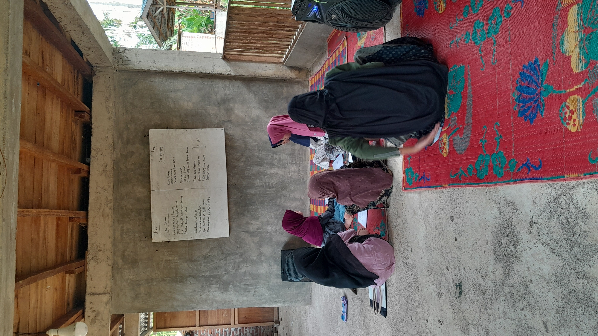 Certainly, let's describe the image.
**Scene:**
The image appears to be taken indoors, likely in a community center or learning space in Aluerambee, Indonesia. The interior features a cement/concrete wall, wooden beams, and a basic structure.
**People:**
There are four individuals, all women, seated on the floor on colorful mats. They are wearing headscarves in various colors. They are facing a whiteboard. They are bent over and appearing to be reading and/or writing in notebooks.
**Objects:**
1. **Whiteboard:** A whiteboard with handwritten text in Indonesian is mounted on the wall. One section is titled "Puisi. Cita-Citaku" (Poem. My Aspiration) and another section is titled "Lagu. Stop Bullying" (Song. Stop Bullying).
2. **Mats:** Colorful plastic woven mats serve as seating on the floor.
3. **Notebooks:** Each woman has a notebook in front of them.
4. **Speaker:** A black speaker is visible on the floor.
**Actions/Interactions:**
The women appear to be engaged in a learning activity, reading and possibly copying information from the whiteboard. They are seated in a focused and attentive manner.
**Environment:**
The setting seems to be a simple, functional space used for educational or community activities. The lighting seems adequate but the interior is basic. The focus is on the people and their learning activity.