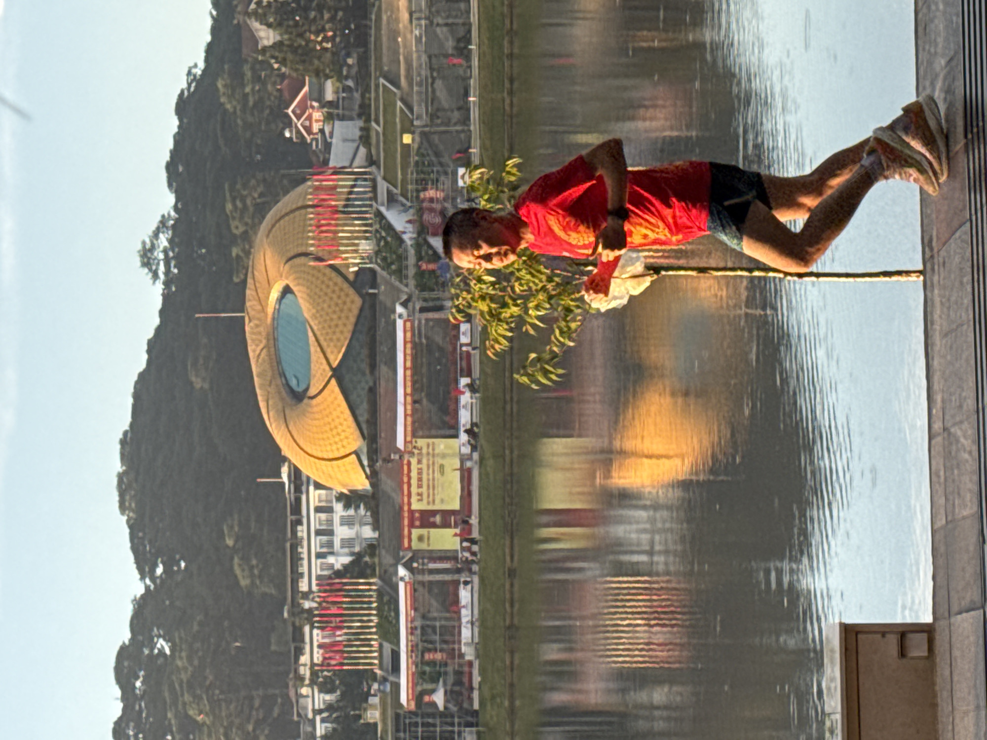 A man is running in the foreground of the image, wearing a red t-shirt and blue shorts. He is holding a white object in his left hand and a stick in his right hand. Behind him is a body of water, likely Xuan Huong Lake, with a reflection of the buildings and trees on the opposite side. In the background, there is a distinctive golden building with a circular opening, which is the Da Lat Flower Garden or the Da Lat Exhibition Center. The area appears to be a public space in Da Lat city, Vietnam, given the architecture and vegetation. The time of day appears to be either sunrise or sunset, as indicated by the lighting and shadows.