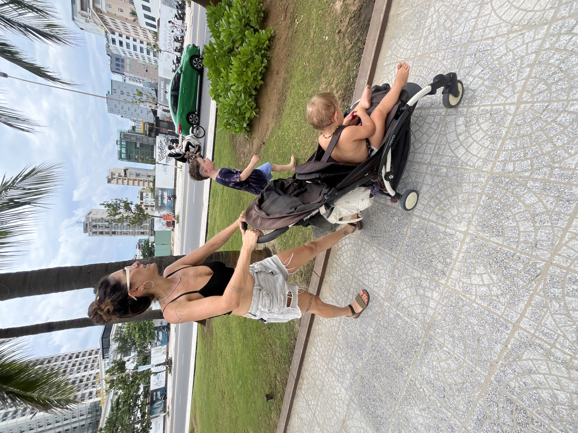 In Đà Nẵng, Vietnam, a woman is pushing a stroller along a tiled sidewalk adjacent to a grassy area. A young boy walks in front of the stroller, and a baby is seated inside. Palm trees and urban buildings line the background, with a green car and other vehicles visible on the street. The scene appears to be a public walkway in a city with a coastal feel.