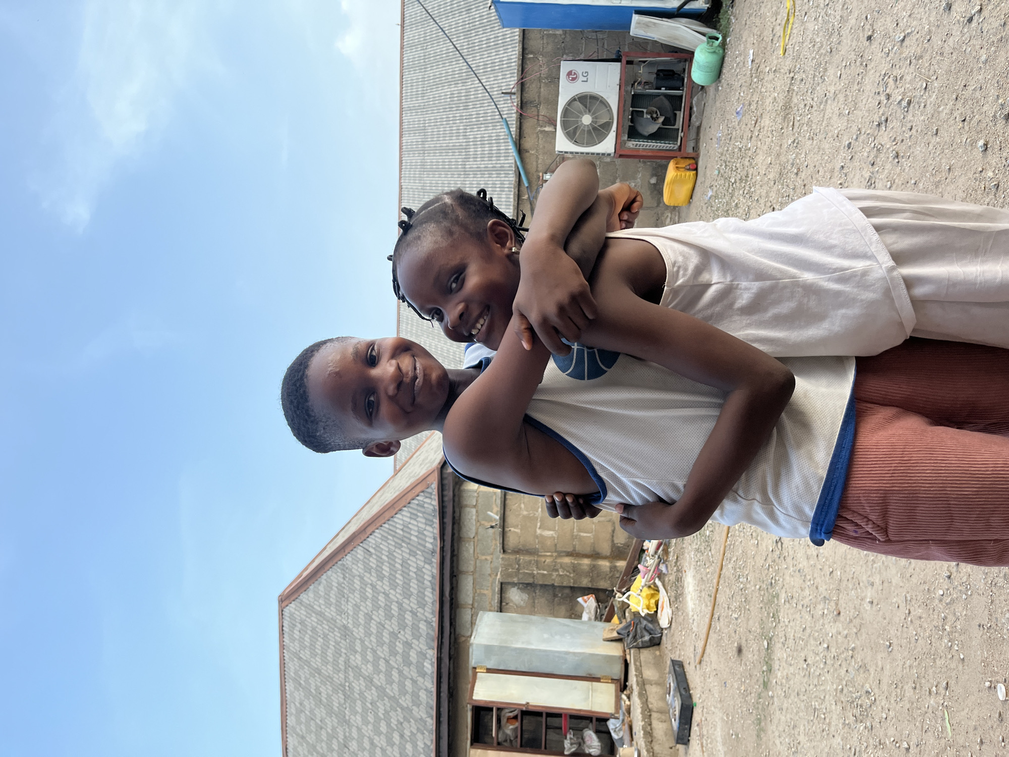 Two children, a boy and a girl, are embracing tightly. The boy is wearing a white tank top with blue trim and rust-colored corduroy pants. The girl is wearing a white top. An air conditioning unit is visible on a building in the background. The setting appears to be an outdoor area, likely in Iddah, Nigeria. The ground is dirt and gravel. Various objects, including a yellow jerrycan and metal containers, are scattered around. The sky is visible above.