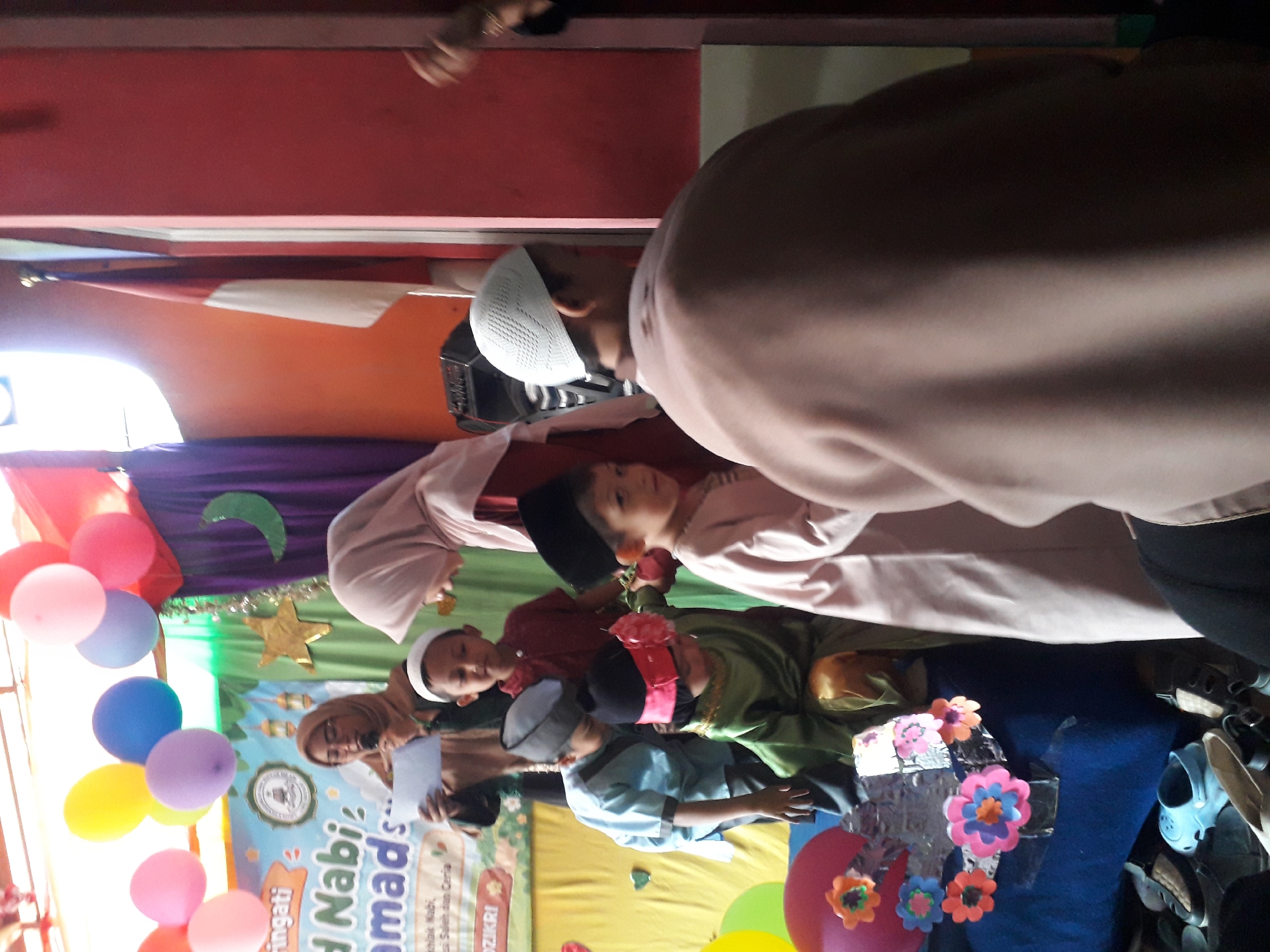 The image captures a lively event, likely a school celebration, at an unspecified location in Cankuang, Indonesia. The foreground is dominated by a group of children dressed in traditional Islamic attire. Several children are wearing peci hats, some in black and others in white. One child in the center is dressed in a green tunic and golden pants, with a red flower adorning their headwear. Another child, wearing a pink headscarf, is visible on the right side of the frame.
Behind the children, an adult woman in a light-colored hijab is holding a piece of paper and speaking into a microphone, suggesting she might be a teacher or speaker. Balloons in various colors, including pink, yellow, and blue, are suspended in the background, adding to the festive atmosphere. A banner with Indonesian writing is also visible, and a green crescent moon decoration is hanging on a purple curtain.
The environment appears to be an indoor space, possibly a community hall or classroom, with a stage area decorated for the occasion. The lighting suggests it's daytime, with some natural light filtering in from the left. The overall impression is one of a cultural and religious gathering, celebrating an important event.