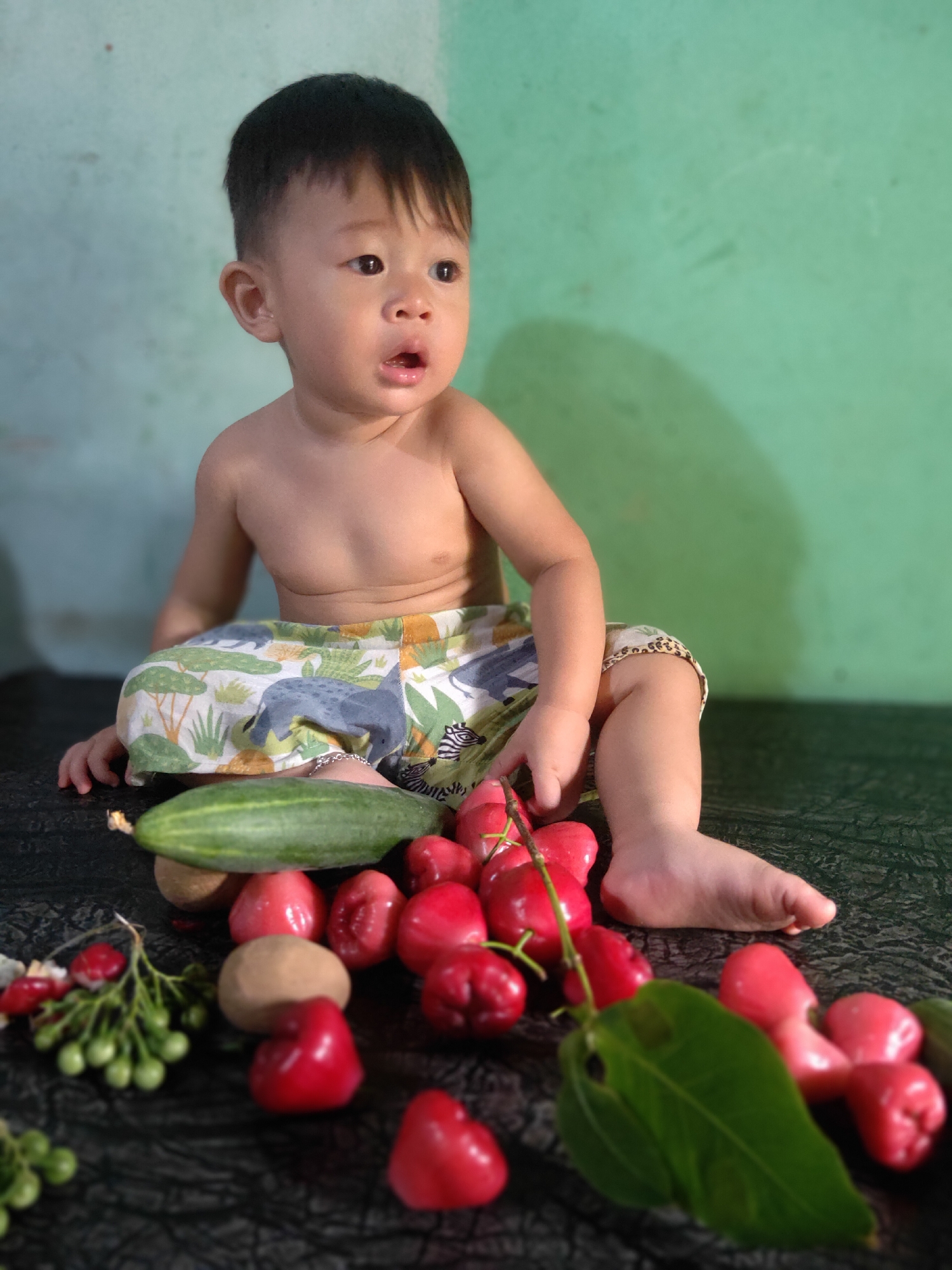 A shirtless young child, seated, is surrounded by various fruits and a cucumber on a dark, textured surface. The child wears shorts with a patterned print featuring animals. The background is a plain, light green wall. The scene is likely indoors, given the lighting and background.