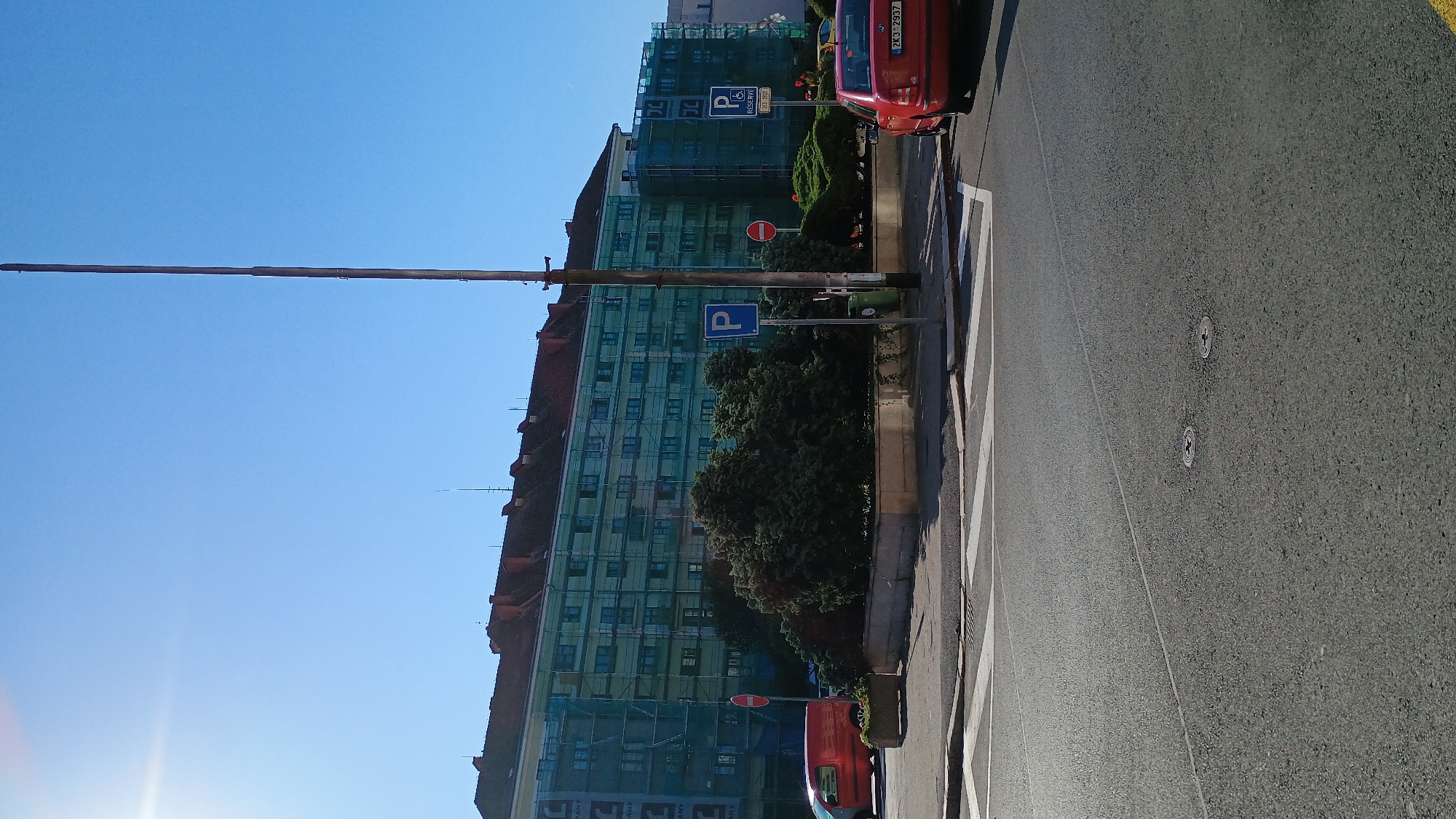 The image shows a street scene in Sokolov, Czechia. In the foreground is a paved road with white crosswalk markings. Two round metal covers are visible on the road surface. In the middle ground, there is a raised planter with dense green foliage. Beyond the planter is a large building covered in scaffolding and green netting, suggesting renovation or construction. Several parking signs, including one marked with a blue "P" for parking and another for disabled parking, are visible. A red van is parked on the left side of the image, and a red car is parked on the right. The sky is clear and blue, with the sun casting a bright glare on the left side of the image. A tall wooden pole with some wires or antennas extends vertically into the sky on the right side of the image. The overall scene depicts an urban environment on a sunny day.
