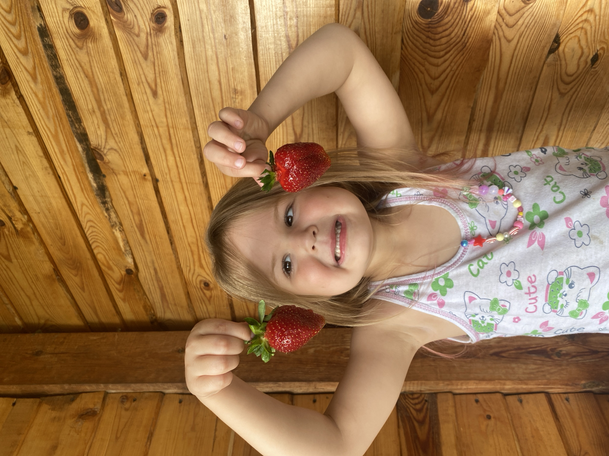 A young girl with blonde hair and pink streaks stands in front of a wooden wall, holding two strawberries. She is wearing a white tank top with a pattern of cats and flowers, and a beaded necklace. Her mouth is open in a smile, and she is looking slightly to her left. The background is composed of horizontal wooden planks. The location is not specified in the image.
