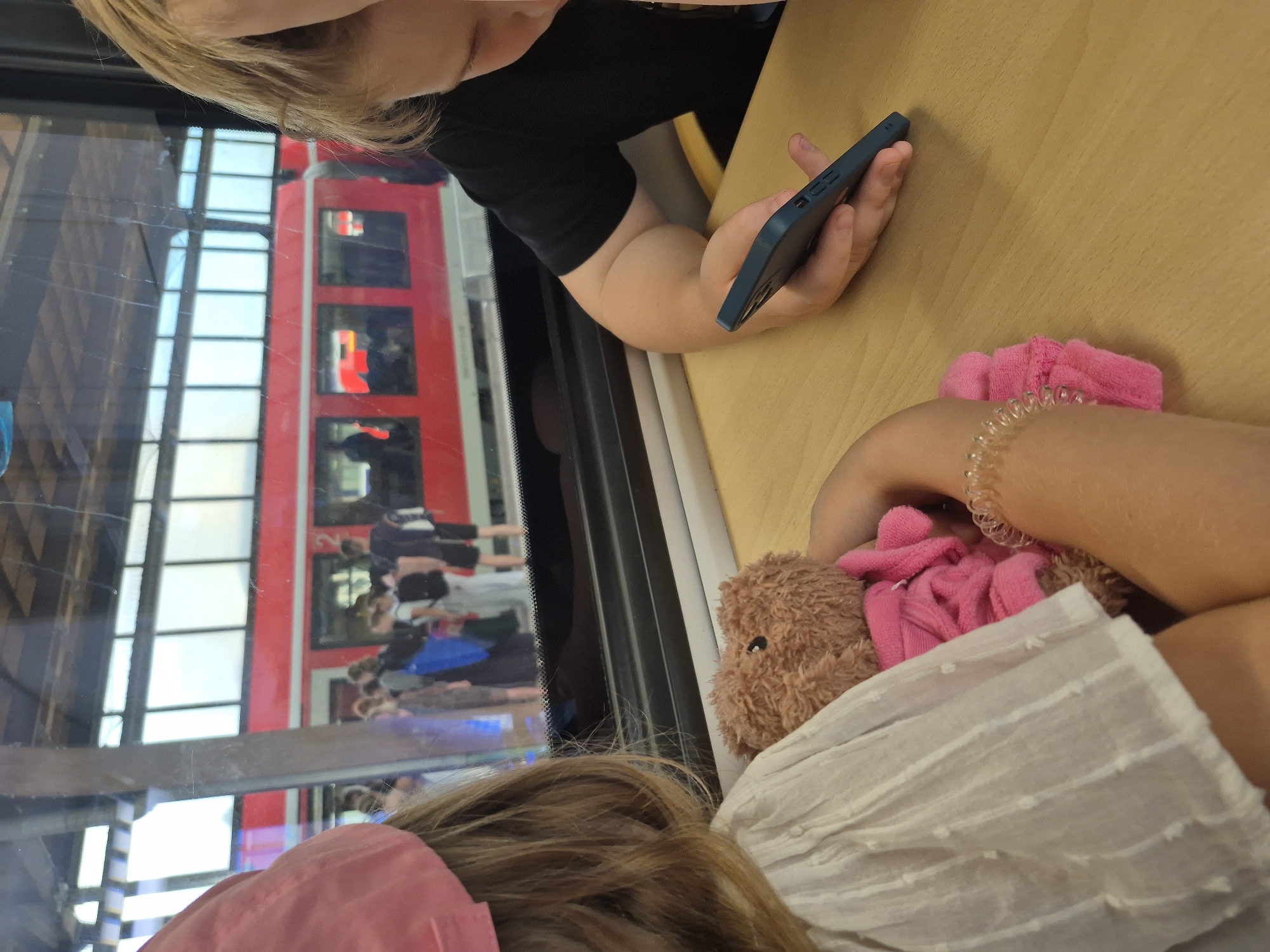 Two children are seated at a table inside a train, looking out of a window. A red train is visible outside the window, stopped at a station platform where several people are standing. The child on the left is wearing a pink cap and a white dress and is holding a teddy bear. The child on the right is wearing a black shirt and is looking at a smartphone. The scene appears to be at the Trier Hauptbahnhof (central train station) in Trier, Germany, given the distinctive red and white German train design and the architecture of the station visible through the window.