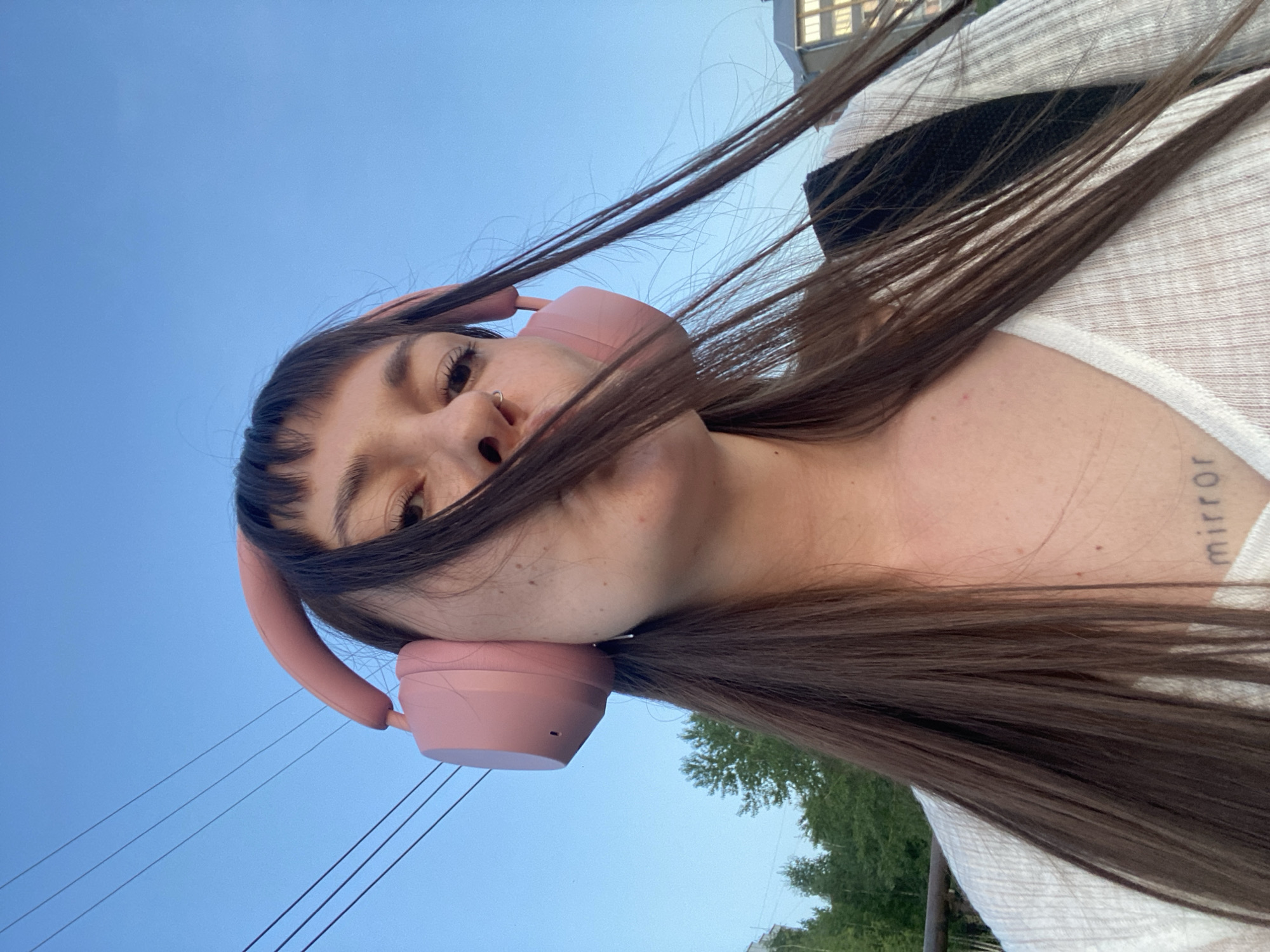 A low-angle perspective captures a woman wearing pink headphones, positioned against a backdrop of a clear blue sky and a tree. The woman has brown hair and a visible tattoo on her collarbone that reads “mirror”. Power lines are visible in the lower left corner. The location is potentially the city of Votkinsk, Russia.