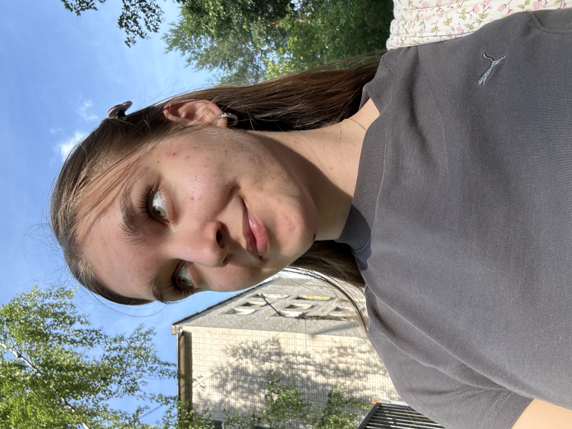 The media file is a portrait-oriented selfie taken outdoors on a sunny day. The subject is a young person with long brown hair, looking towards the camera with a slight smile. They are wearing a dark gray t-shirt with a subtle, light blue embroidered logo on the left side of the chest, which appears to be a puma. The background consists of a clear blue sky with some scattered white clouds, green foliage from trees, and a building with light-colored siding and multiple windows. A decorative patterned fabric with pink flowers and green leaves is visible at the top of the frame, suggesting the person may be leaning against something or lying down. The lighting indicates it is daytime, likely late morning or afternoon, with the sun casting shadows. One of the provided crops shows a small object, possibly a hairpin or accessory, clipped to the subject's hair near their ear. There is no visible text in the image. The location context is Chaikovsky, Russia.