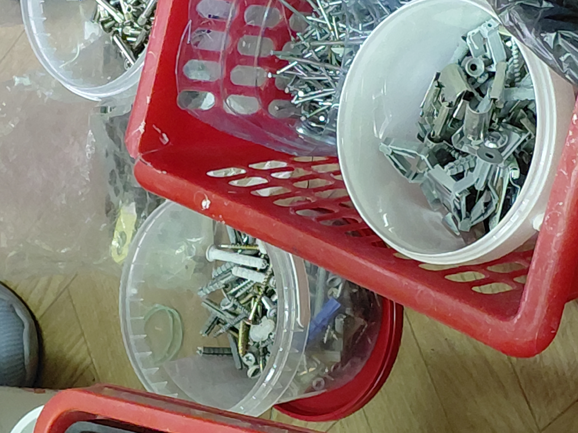 A high-angle shot shows red plastic baskets containing containers filled with various hardware items (screws, nails, anchors, etc.). The location, Nadym, Russia, is not discernible from the image. The composition emphasizes the organization and categorization of fasteners. There are no people visible.