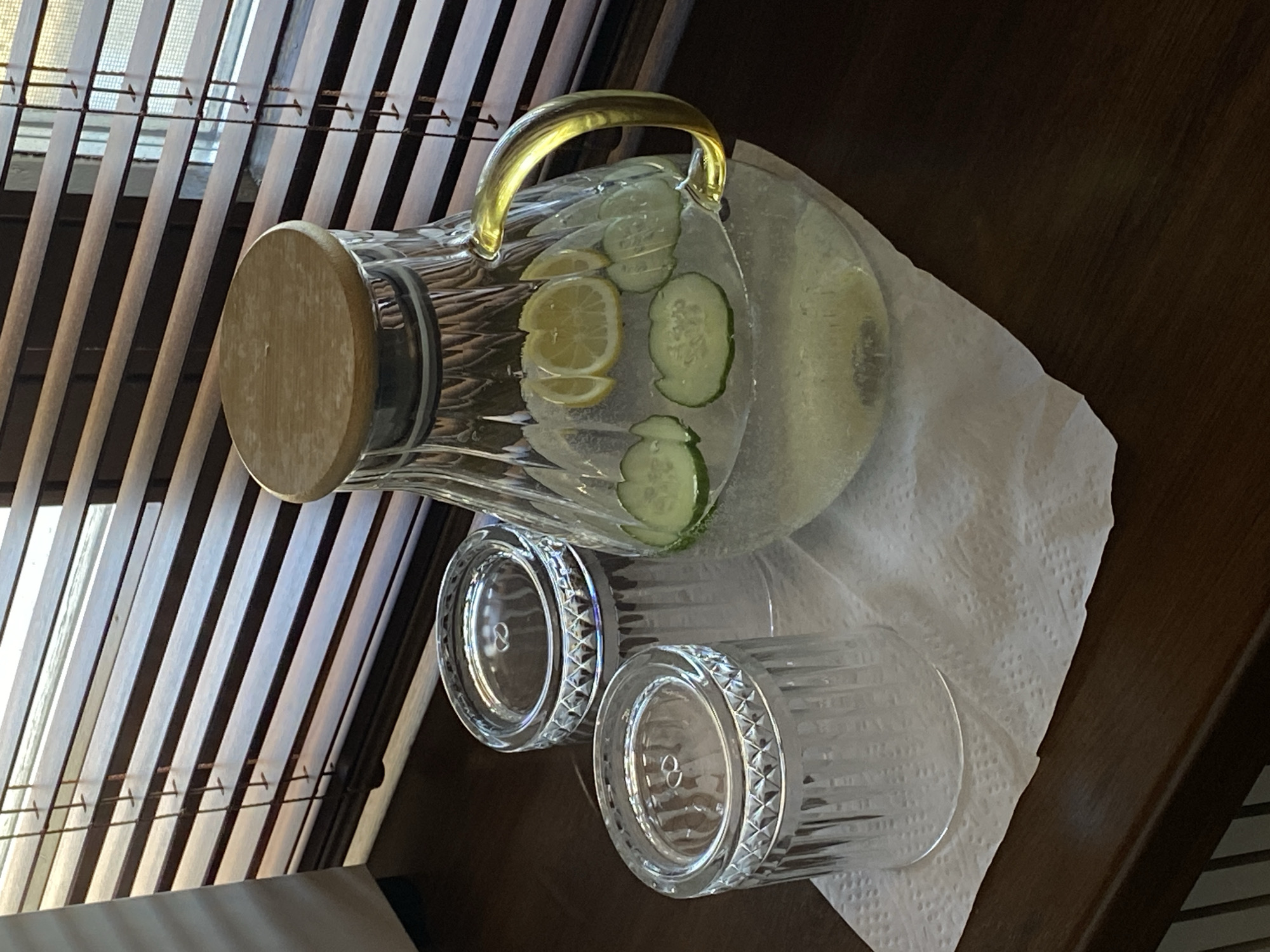 A still life presents a clear glass pitcher filled with water, lemon slices, and cucumber slices, alongside two matching clear glass tumblers, all arranged on a white textured napkin atop a dark brown wooden surface. The pitcher features a golden-colored handle and a wooden lid. The tumblers exhibit a textured design around their bases. A window with closed horizontal blinds is visible in the background, partially obscured by the objects. The lighting is bright, casting shadows that define the contours of the objects and the surface. The precise location cannot be determined from this image, and there is no evidence to suggest it is in Votkinsk, Russia. There are no people depicted in the image, and no actions or interactions are taking place. The environment consists of an interior setting with basic furniture and window coverings.
