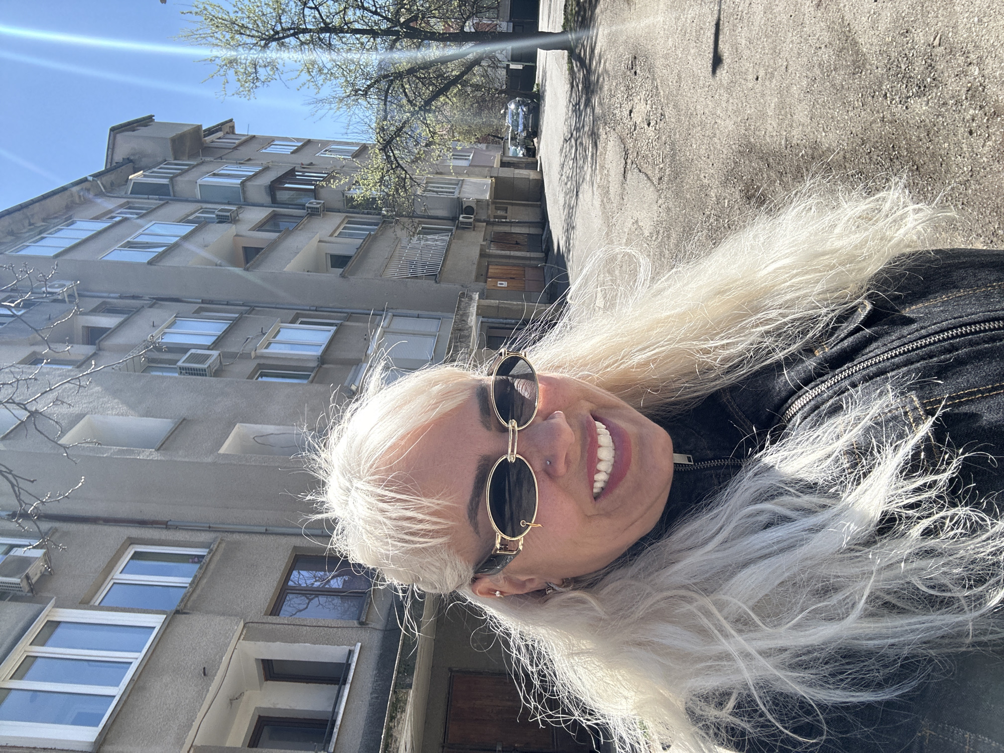 A person with long blonde hair, sunglasses, and a nose piercing is smiling at the camera. They are wearing a dark jacket. In the background is a multi-story residential building with visible air conditioning units and windows. The building is located on a street that appears to be made of asphalt. The weather is sunny and there are trees with sparse foliage overhead. The image was likely taken in Sofia, Bulgaria, given the architectural style of the building.