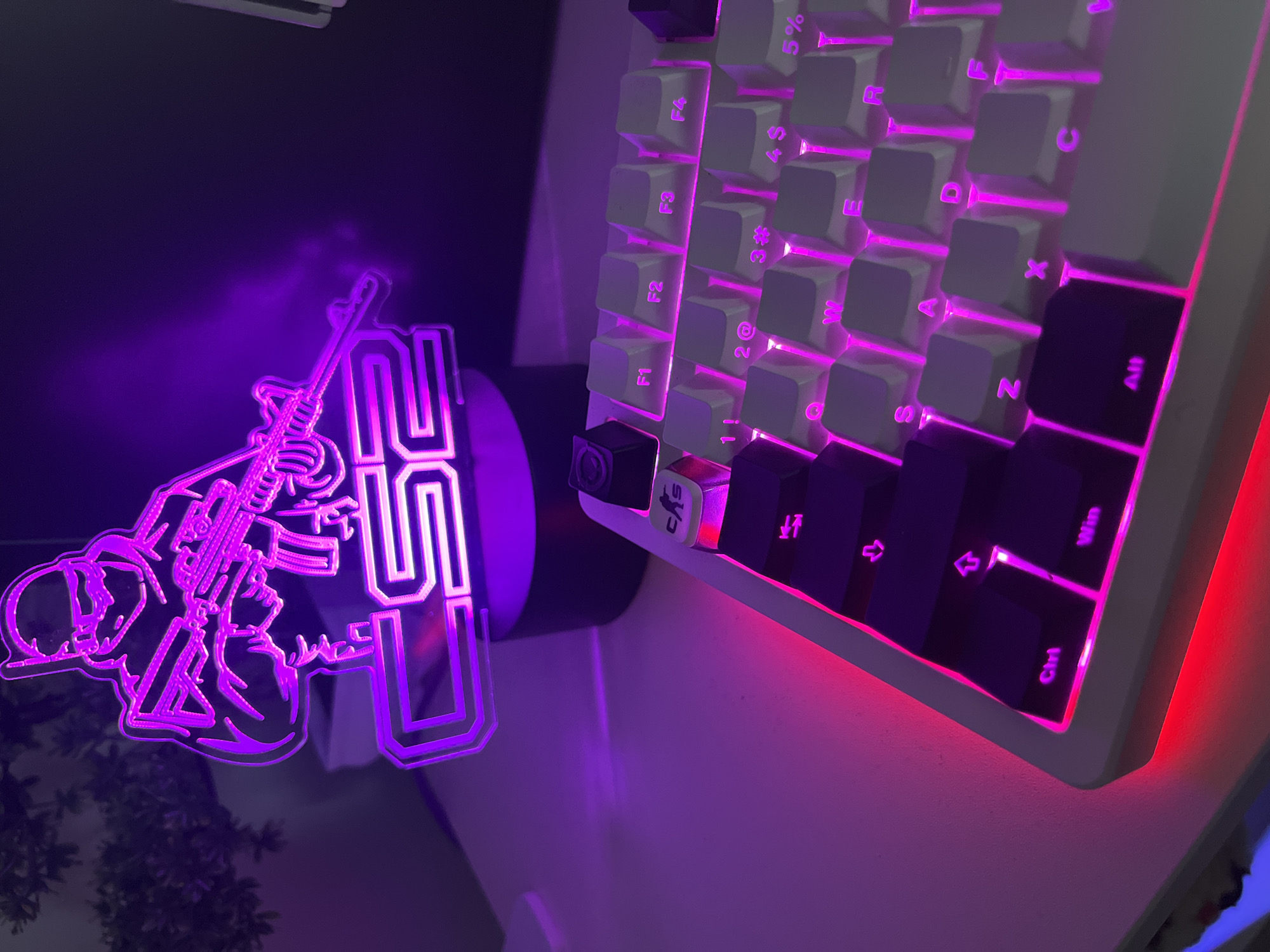 The image depicts a close-up, eye-level view of a white mechanical keyboard with pink backlighting. The keyboard is positioned on a white desk, and a purple LED lamp featuring a silhouette of a character holding a rifle and the text "CS2" is visible in the background. The lamp is casting a purple glow on the desk and keyboard. The keyboard shows several keys, including "Ctrl", "Win", "Alt", and function keys "F1" through "F4", along with alphabetical keys like "Q", "W", "E", "R", "S", "A", "D", "F", "Z", "X", and "C". The overall aesthetic suggests a gaming setup. No specific location in Varna, Bulgaria can be determined from the image.
