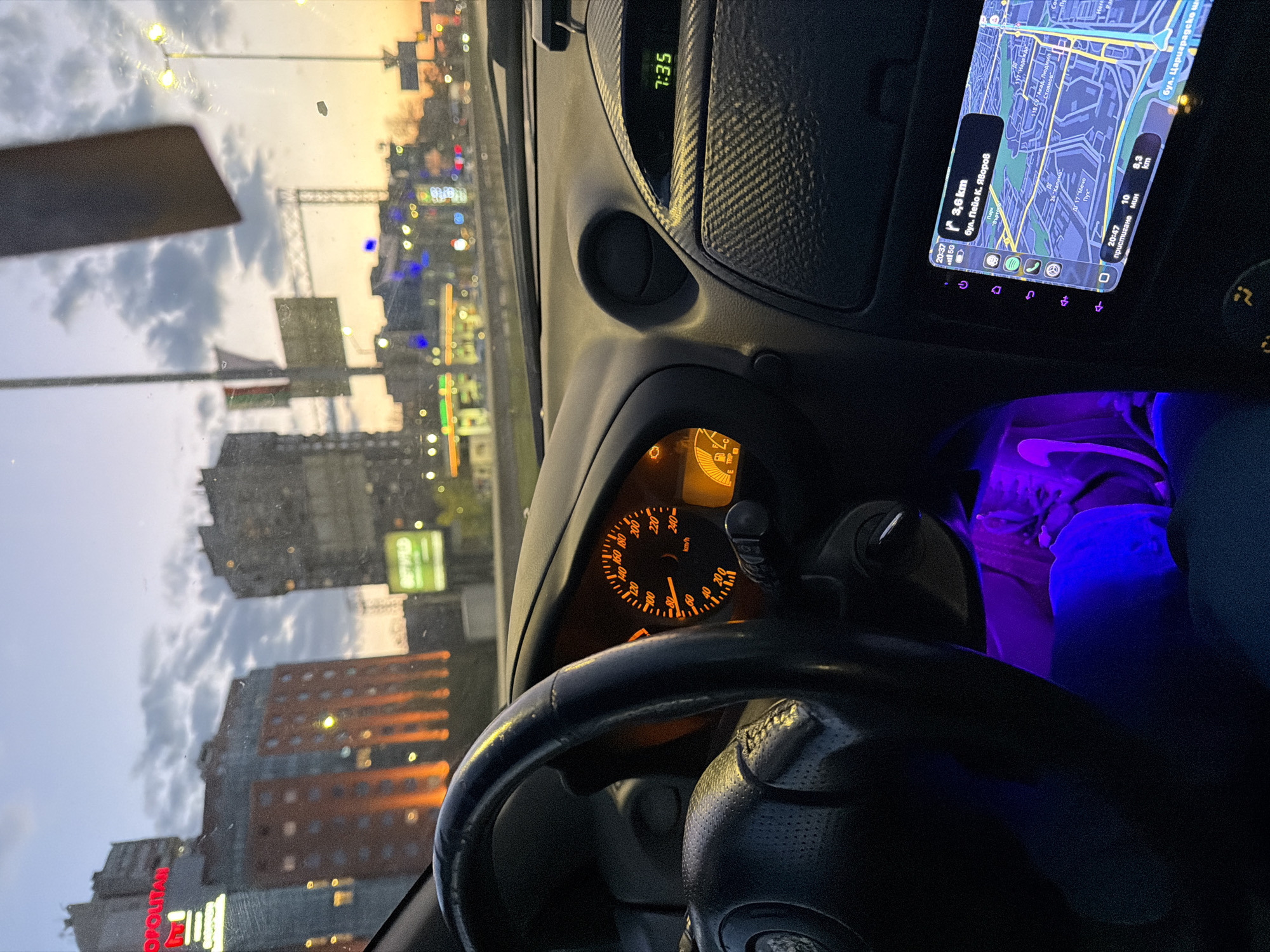 The image captures the interior of a car looking out onto a city street at dusk or early night. The primary subjects are the car's dashboard, steering wheel, and an infotainment system screen, along with blurred buildings and lights outside.
Inside the car, a black steering wheel is prominent in the foreground. Behind it, the instrument cluster shows an orange-lit speedometer at 0 km/h, indicating the car is stationary. A "check engine" light is visibly illuminated. Above the speedometer, a digital clock displays "7:35". To the right, the dashboard features a textured carbon fiber-like trim and an air vent. Purple ambient lighting illuminates the footwell, where a person's leg is partially visible.
The infotainment screen, displaying a navigation map, is a central feature. The map shows street names in Cyrillic, including "бул. Пейо К. Яворов" (Peyo K. Yavorov Boulevard) and "бул. Цариградско шосе" (Tsarigradsko Shose Boulevard), confirming the location as Sofia, Bulgaria. The screen indicates the current time as "20:37" (8:37 PM), with an estimated arrival time of "20:47" (8:47 PM) for a destination 8.3 km away. Another destination 3.6 km away is also shown. A Spotify icon is visible among other app shortcuts at the bottom of the screen.
Outside the car, through the windshield, the sky is cloudy with residual light from the setting sun, transitioning into night. City lights from buildings, streetlights, and traffic are visible. Partially visible signs on buildings include red letters that seem to spell "METROPOLITAN" and a green sign with "SCTHD". The overall scene suggests a typical urban environment during the evening commute, with the car either parked or stopped in traffic.