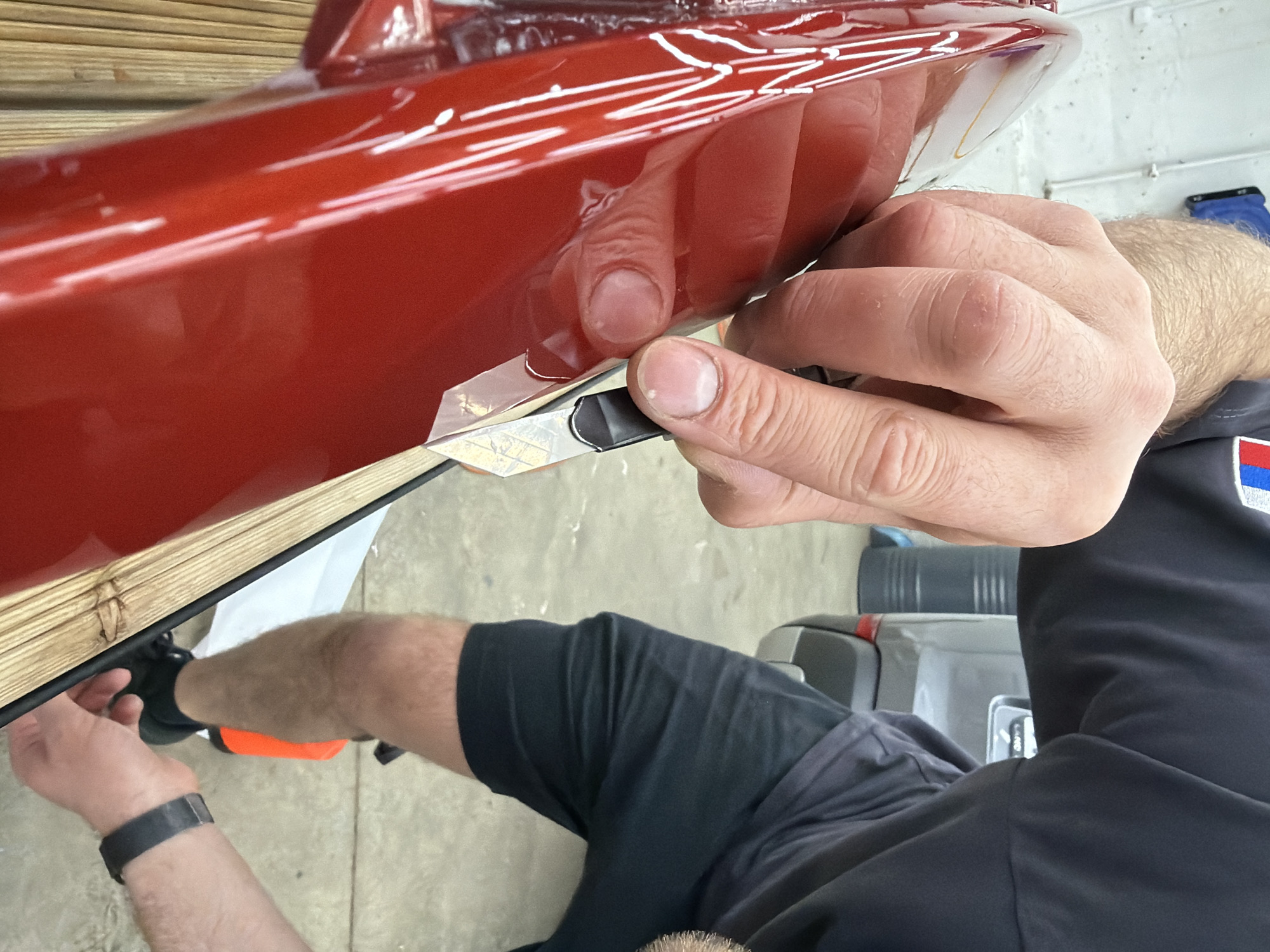 This image depicts an indoor workshop or garage environment. A male individual, largely seen from the mid-torso down and his right arm, is actively working on a glossy, highly reflective, deep red object, consistent with a vehicle body panel. He wears a dark, short-sleeved shirt with a small, multi-colored emblem on the right sleeve, black shorts, black athletic shoes, and black socks. His right hand, positioned prominently in the foreground, is holding a utility knife with a silver blade and a black handle, precisely trimming or scoring a transparent film applied to the red surface.
A second individual's left hand, adorned with a black wristband, is visible lower in the frame, near the base of the red object and a light-colored, slatted wooden support structure upon which the red object rests.
In the background, a grey SUV or car is partially visible, showing its rear section, including a red taillight and an area where a license plate would be mounted, though none is present. Faint lettering, possibly "CAND," is discernible on the rear of the vehicle. To the left of the vehicle, a dark grey or black barrel-shaped container is present. The floor is light-colored concrete. An orange object, potentially a tool or bottle, and a white sheet are visible on the floor near the grey vehicle. The walls are light-colored, consistent with an industrial or service setting.
The specific location within the city of Krasnoyarsk, Russia, cannot be determined from the visual information provided in the image.