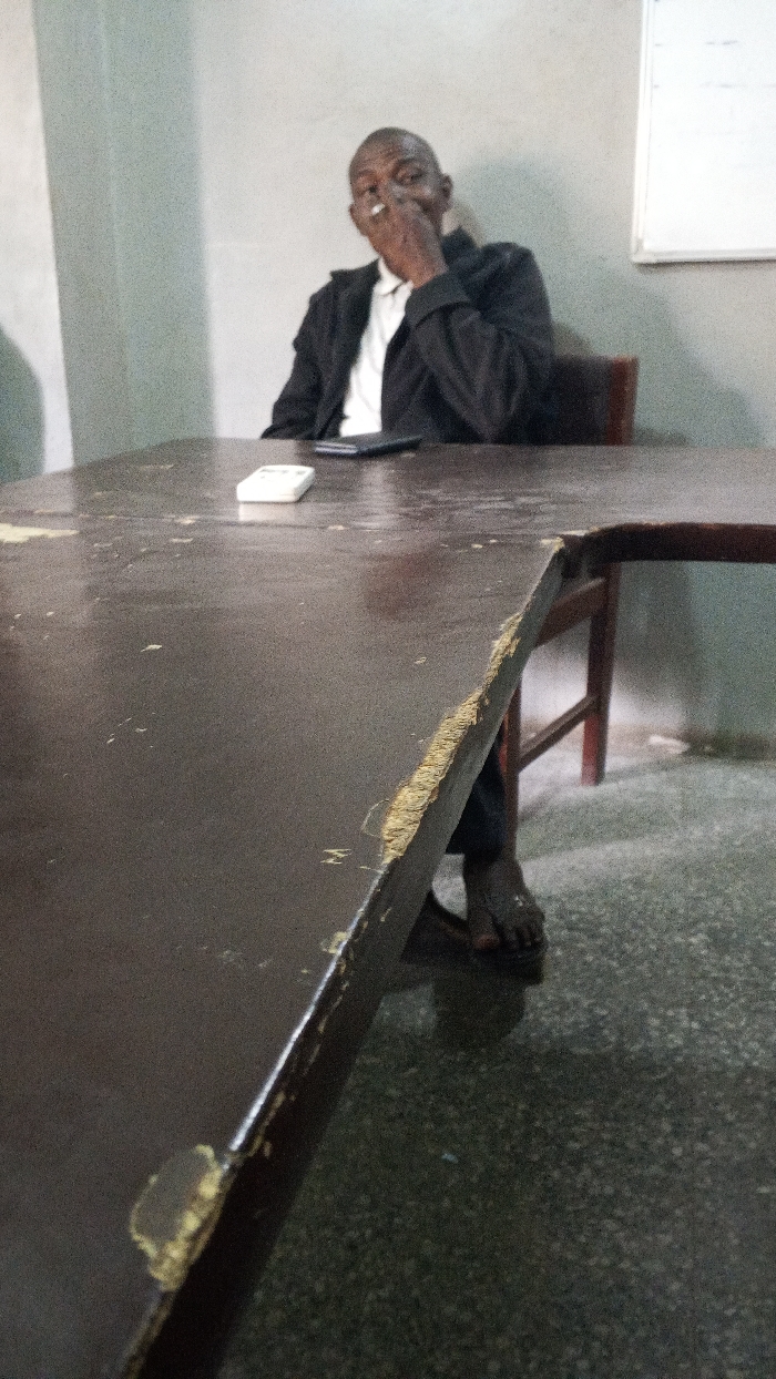 The scene is an indoor environment in Dakata, Nigeria. An adult male with dark skin is seated at a dark brown, heavily worn table. He is dressed in a white collared shirt and a dark blazer. His right hand is raised to his face, holding a white cylindrical object, consistent with a cigarette, between his fingers. A white rectangular object, resembling a remote control, and a dark smartphone are positioned on the table surface in the foreground. The table shows significant wear, with multiple chips, scratches, and areas where the laminate is peeling, exposing lighter material beneath. The man's left foot, bare and wearing a dark sandal, is visible under the table. The background features plain, light-colored walls and a dark, speckled floor. A white rectangular board is visible on the wall to the right of the man.