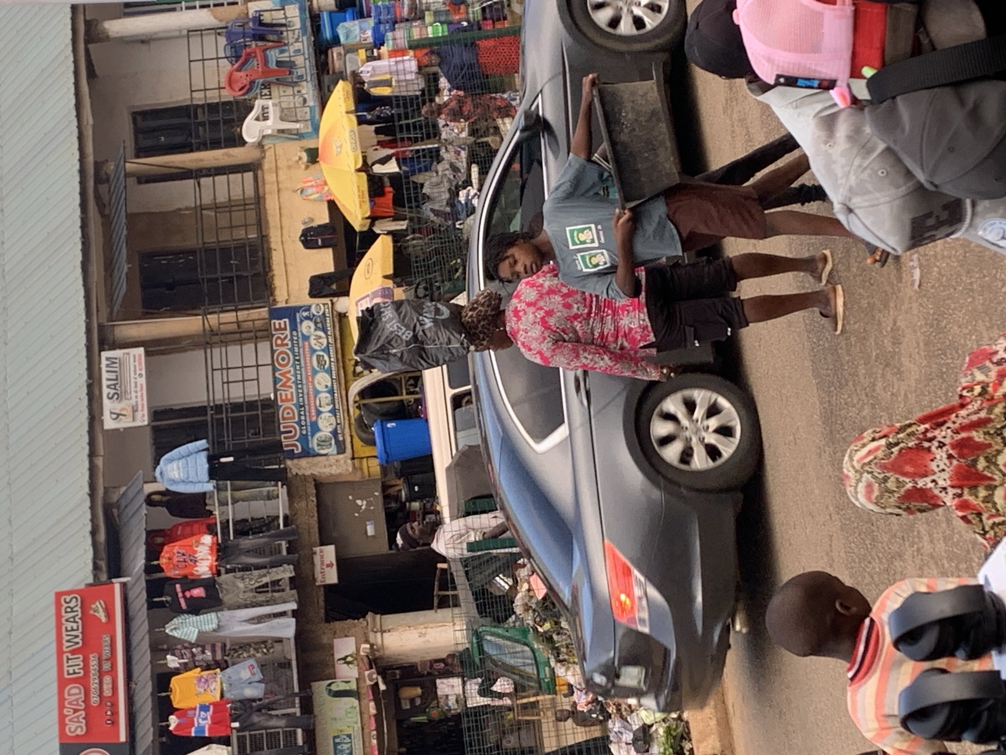 This is a medium shot of a busy market scene in Bukuru, Nigeria. The image is oriented vertically, with the ground occupying the bottom third and buildings lining the left side. A dark-colored car is parked in the middle of the frame, partially obstructing the view of the market stalls.
Several people are visible. In the foreground on the right, a young child wearing a striped sweater is looking away from the camera. To the left of the child, a person in a red and white patterned top is partially visible. In the center of the frame, two individuals are interacting near the car. One person, wearing a pink and white floral top and a leopard-print headscarf, is looking down. Beside them, another person in a blue t-shirt and dark shorts is holding a black tray. Further in the background, more people are seen among the market stalls.
The market appears to be selling clothing and other goods. Numerous items are displayed on racks and shelves, with some clothing items hanging outside the shops. Several umbrellas, some yellow, are also visible, suggesting it might be a sunny day, or they are for shade. Signs with text like "SAYAD FIT WEARS," "SALIM," and "JUDEMORE GLOBAL INVESTMENT LIMITED" are present on the buildings, indicating the names of businesses. An "Entrance" sign is also visible. The overall impression is one of a bustling marketplace with a lot of activity and merchandise. The lighting suggests it could be daytime.