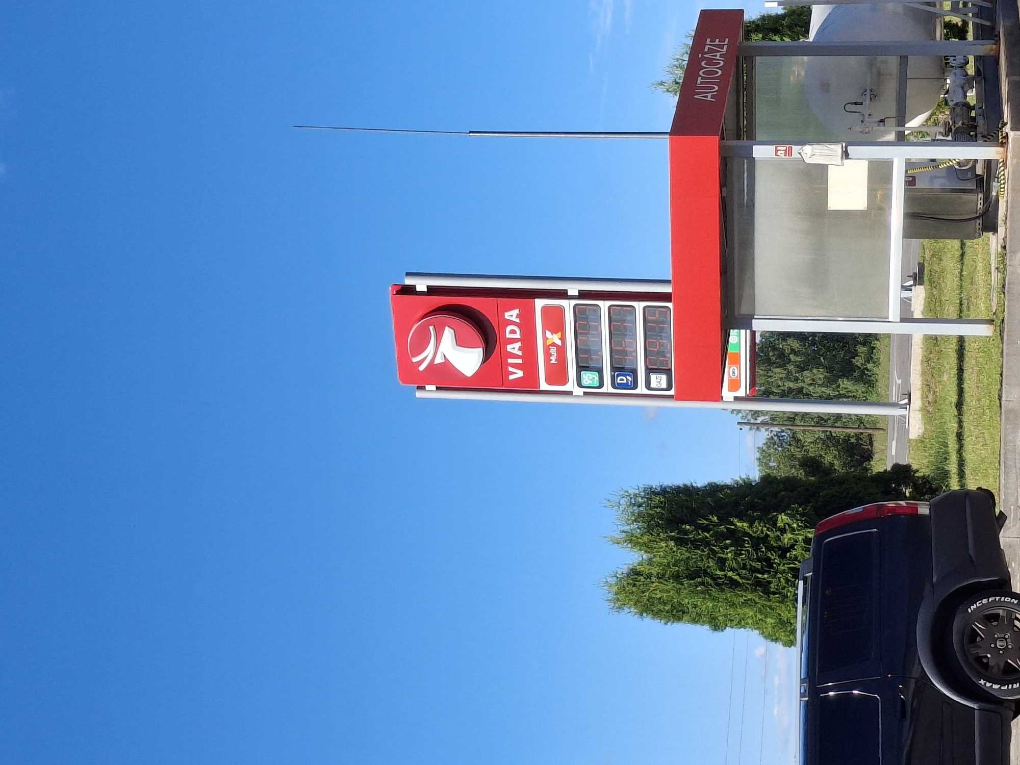 This media file captures a bright, sunny day in Glāžšķūnis, Latvia, with a clear blue sky dominating the scene. The primary focus is a red gas station sign displaying fuel prices. The sign prominently features the "VIADA" logo and the brand name "VIADA" in large white letters. Below, digital displays show fuel prices for different grades, including "MultiX" and what appears to be "95" octane. Other labels like "D" and "GAZE" are also visible, indicating different fuel types or services.
To the right of the main sign, a section labeled "AUTOGĀZE" suggests the availability of autogas. This area includes a glass enclosure with some equipment visible inside, possibly related to fuel storage or dispensing.
In the foreground, the rear of a dark-colored SUV is visible, parked at the gas station. The tire of the SUV has white lettering on its sidewall, with "INCEPTION" and "RIPAAX" being partially readable. Lush green trees form a backdrop behind the gas station, and a patch of grass is visible along the roadside. The overall atmosphere is one of a typical day at a fueling station, with no people or specific events depicted. The time of day appears to be mid-morning or afternoon due to the strong sunlight and clear sky.