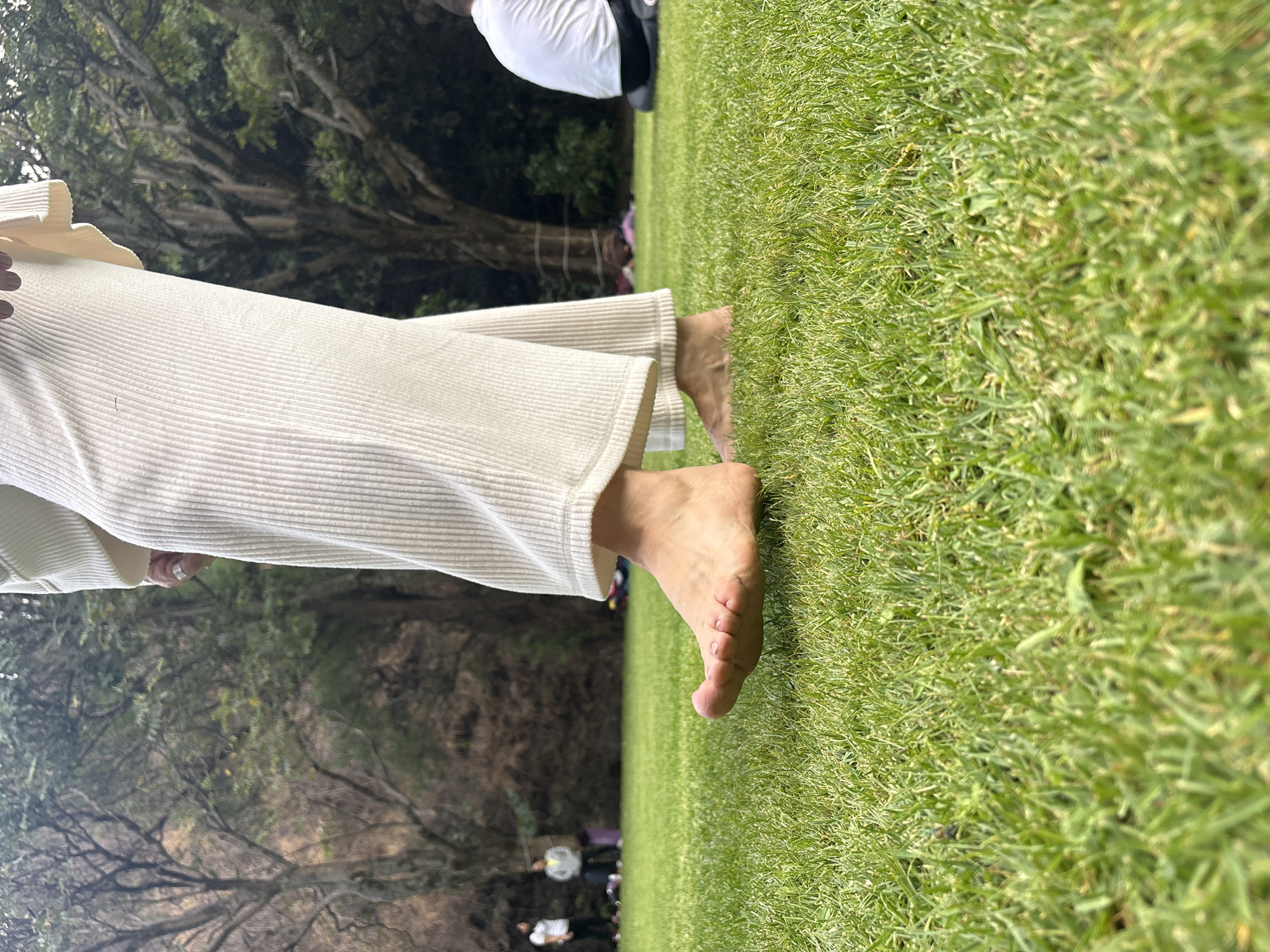 The image shows a person standing barefoot on a grassy area in Santa Ana Acozautla, Mexico. The person is wearing light-colored, corduroy-like pants. The grass is green and appears to be well-maintained. In the background, there are trees and other people, though they are somewhat blurred. The scene seems to be outdoors in a park or natural setting.