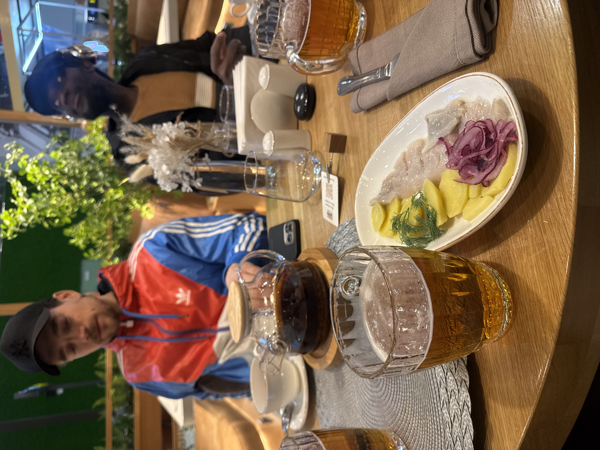 The image captures an indoor dining scene at what appears to be a restaurant or cafe in Ul'yanovka, Russia. Two individuals are seated at a wooden table, engaged in dining and socializing.
In the foreground, a man wearing a distinctive red, blue, and white Adidas track jacket and a black baseball cap is visible on the right side of the table. The Adidas logo (trefoil and three stripes) is clearly visible on his jacket. Across the table and further back, another person is seated, wearing a black top over a light brown or beige striped shirt. Their faces are partially blurred.
The wooden table is laden with various items:
- A white oblong plate holds a dish of sliced herring, boiled potato chunks, pickled red onions, and fresh dill.
- Two large, textured glasses filled with amber liquid, likely beer, one prominently in the foreground with a foamy head.
- A glass teapot containing dark tea, placed on a wooden stand, next to a white teacup and saucer.
- A folded grey cloth napkin with cutlery.
- White paper napkins, salt and pepper shakers, an empty glass, and a small vase with dried white floral arrangements.
- A smartphone is also visible near the teapot.
The setting features warm, ambient lighting. Green foliage, possibly from indoor plants, is visible in the background, adding to the relaxed atmosphere. A small QR code tag is attached to one of the empty glasses on the table.