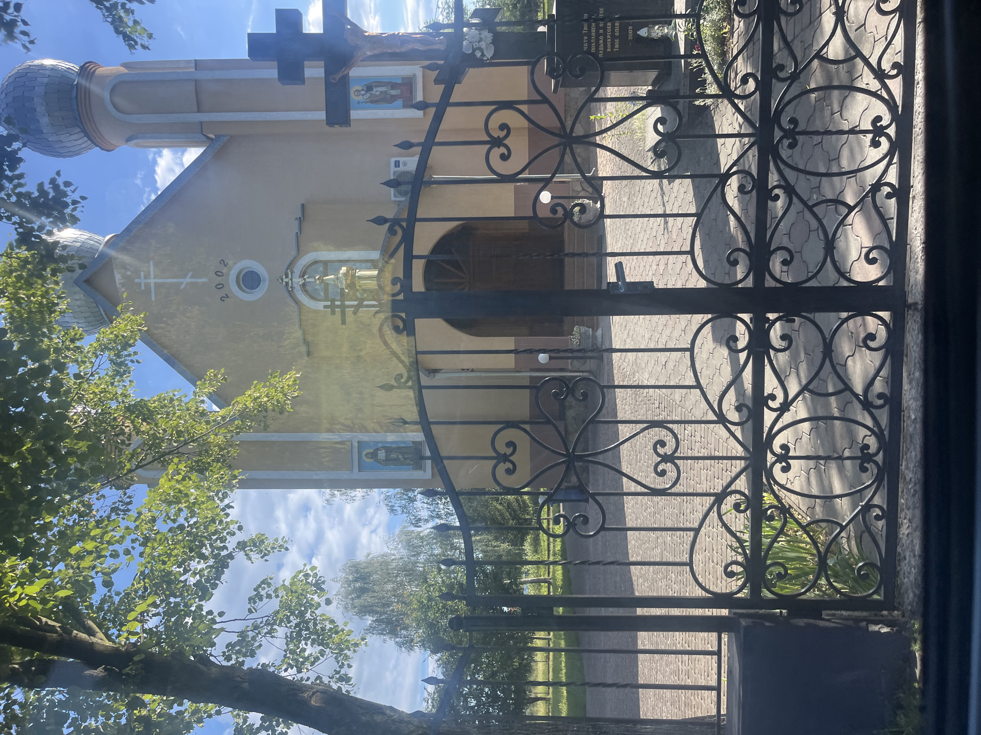 The image shows a view through a decorative black metal gate towards the Church of the Resurrection of Christ in Shyshlivtsi, Ukraine. The church is a light yellow building with two onion domes, one on the main structure and one on a smaller tower. A large black crucifix stands to the right of the church entrance, with a statue of Jesus Christ affixed to it. Above the church entrance, a circular window is visible, and the year "2002" is inscribed on the facade. To the left of the entrance, a painting of a saint is displayed on the exterior wall. The gate is ornate, featuring scrolling metalwork and pointed finials. Behind the gate, a paved pathway leads to the church. Green trees and foliage are visible in the background, and the sky is blue with scattered clouds. A portion of a stone monument with an inscription is visible to the right of the gate. The image appears to have been taken on a sunny day.