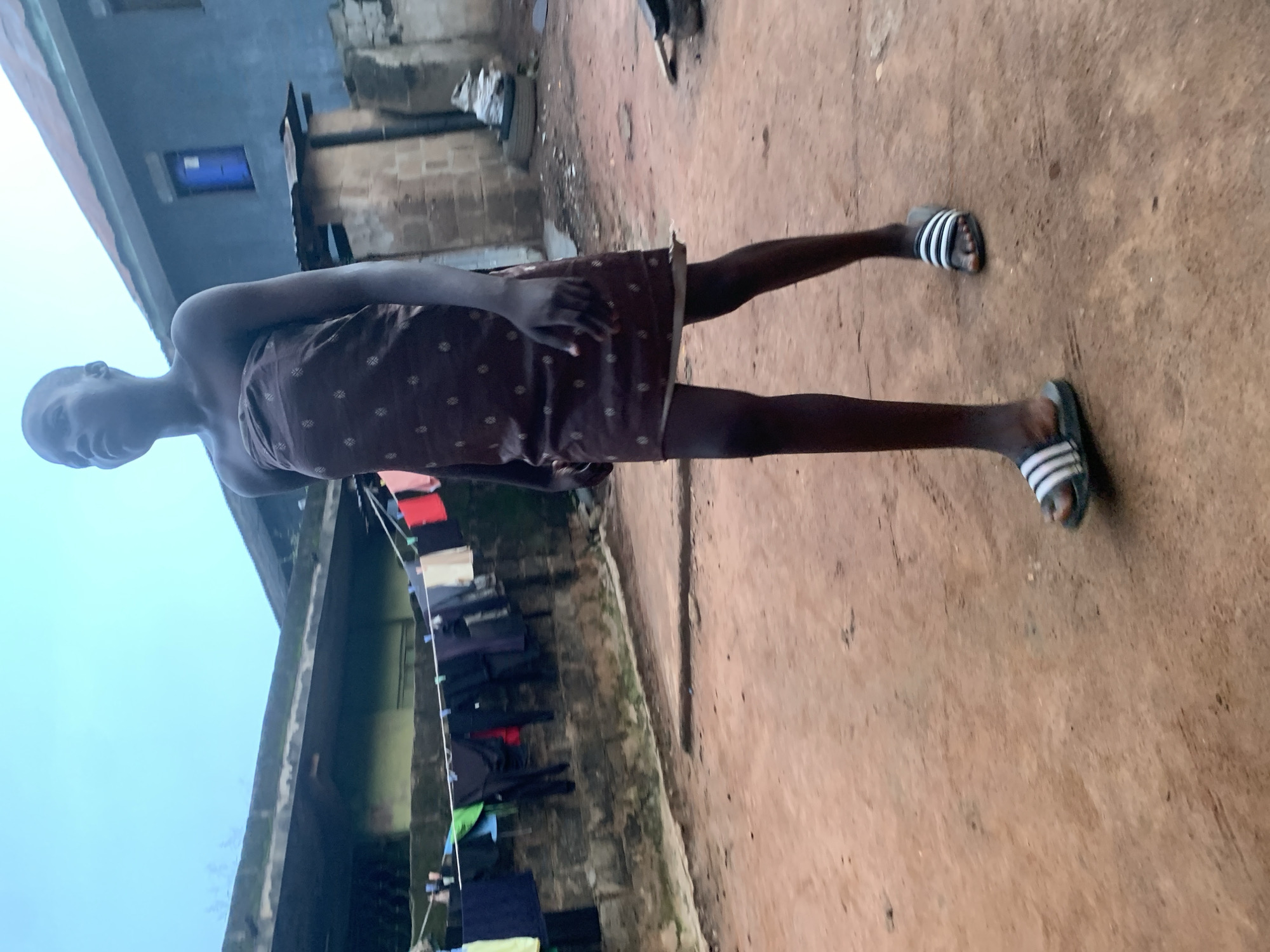 In Shagamu, Nigeria, a young person walks across a dirt path. They are wearing a brown, patterned strapless dress and black and white striped slides. In the background, laundry hangs on a clothesline strung between two buildings. One building is a light green, multi-story structure, and the other is a darker blue building with a blue window. The ground is a reddish-brown dirt, and the sky is a pale blue.