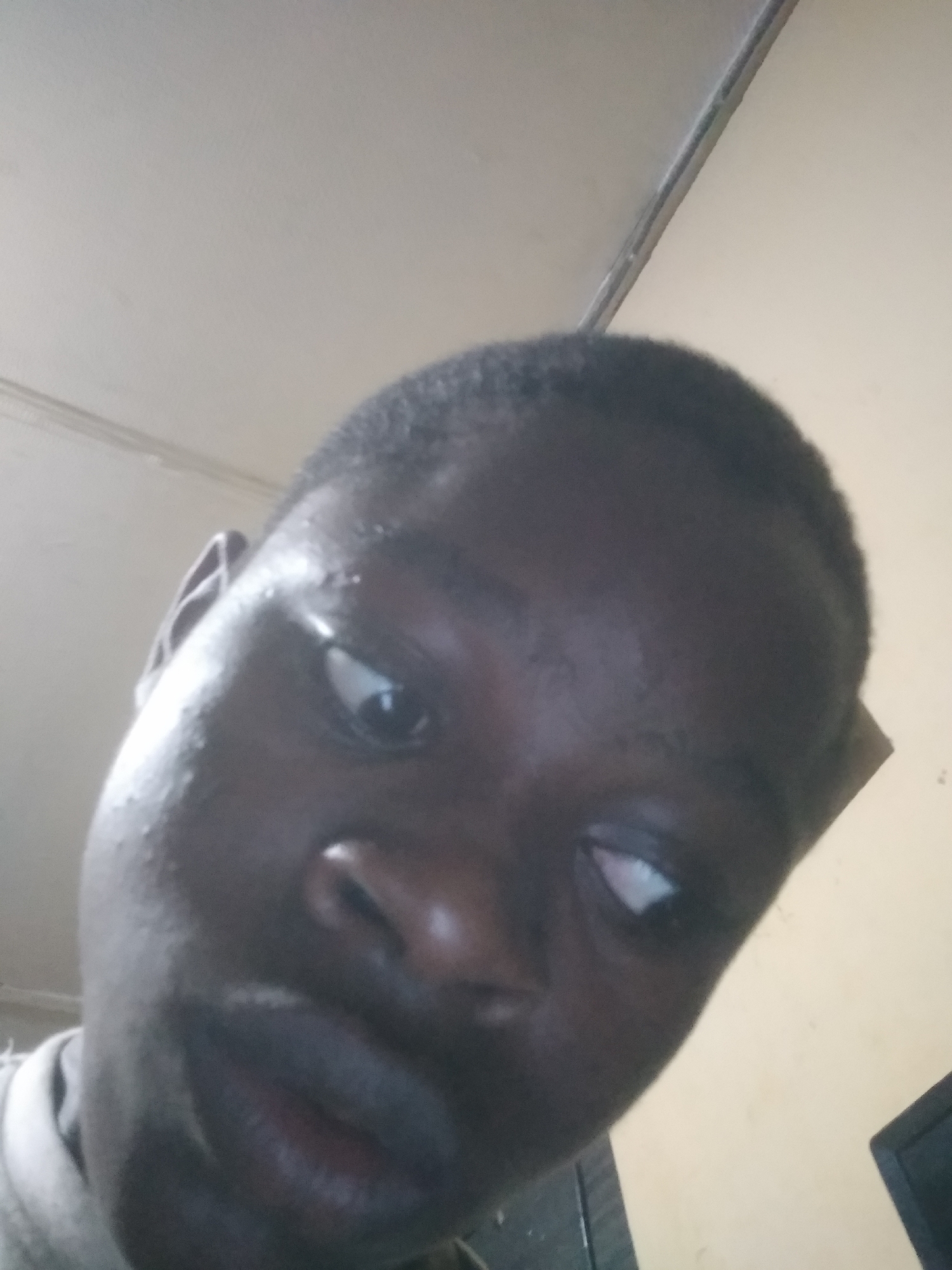 The image is a close-up, low-angle portrait of a young person, likely male, with dark skin and short, dark hair, filling most of the frame. The subject's head is tilted slightly, and their eyes are cast to the right, away from the camera. Light reflects off the forehead and cheek, highlighting some skin texture. The expression is neutral, without strong emotion.
The background is a simple indoor setting featuring a light-colored (off-white or beige) wall and ceiling. A distinct line where the wall and ceiling meet runs diagonally across the upper right corner, with a thin, linear object, possibly a pipe or trim, visible along it. A small, dark object, potentially a monitor or screen, is partially visible at the bottom right edge of the frame. The subject appears to be wearing a light-colored, possibly grey, collared garment, with a small portion visible near their neck and left shoulder. No text is visible in the image. The specific time of day and exact location cannot be determined, other than being an interior space.