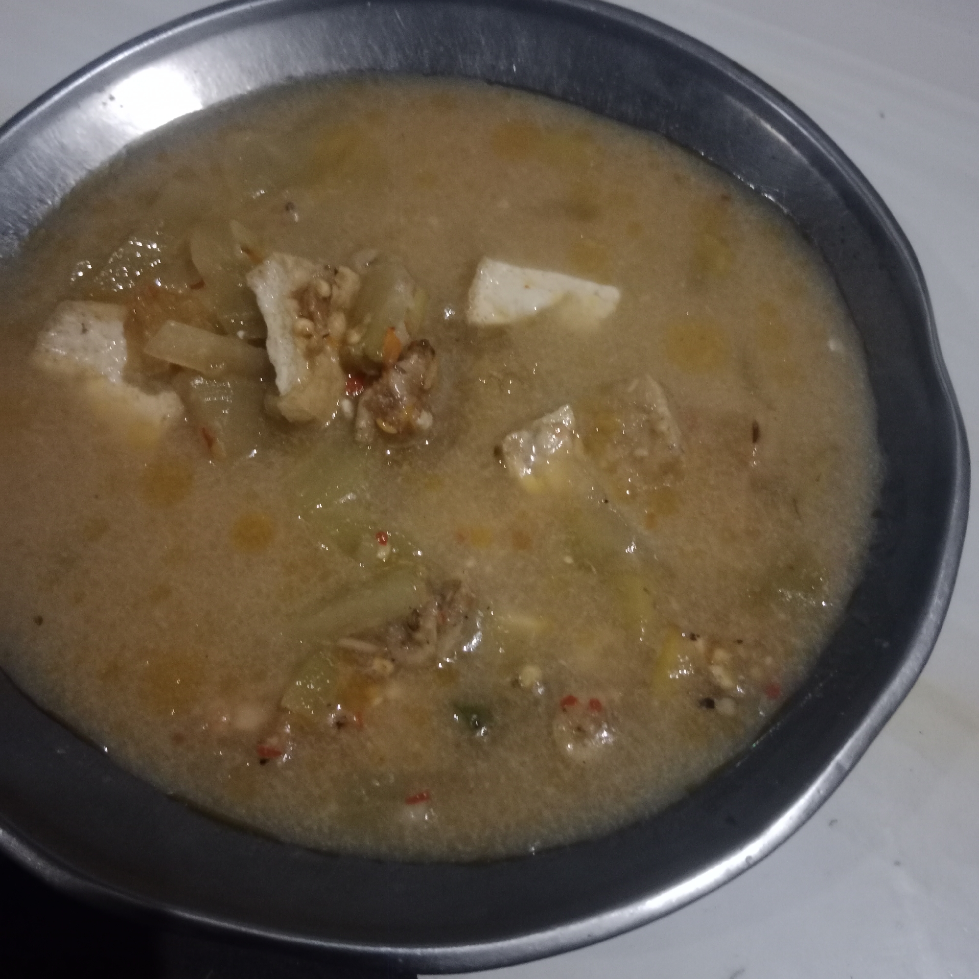 A close-up, overhead shot captures a shallow, metal bowl filled with a light brown, soupy dish. The dish contains visible chunks of what appear to be light-colored vegetables, possibly tofu or gourd, and some darker, possibly meat pieces. Specks of red and green, likely chili or spices, are scattered throughout the broth. The bowl itself has a slightly worn, metallic sheen and rests on a plain, light-colored surface, possibly a table. The overall lighting is dim, creating a soft focus on the food. There are no people or discernible environmental features outside the immediate frame of the bowl. This image is not from Tegowanu Kulon, Indonesia.