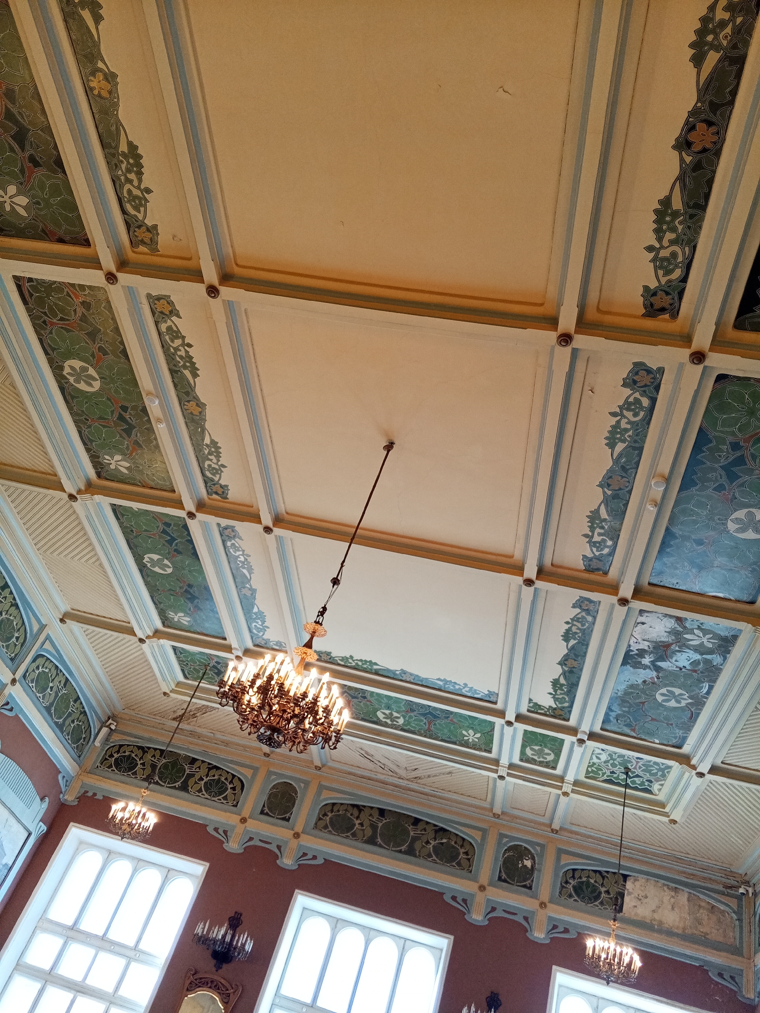 The image displays a detailed, upward-angled view of the interior ceiling and upper walls of the Eliseyev Brothers' Store (Eliseyevsky Gastronom), located at Nevsky Prospekt, 56, Saint Petersburg, Russia.
The high ceiling features a light-colored coffered design, characterized by a grid of beams accented with thin blue and yellow stripes. Within this grid are alternating plain beige rectangular panels and panels containing elaborate green and blue patterns, suggesting stylized floral and organic motifs, possibly executed in stained glass or paint. Small, round, brown decorative elements are situated at the beam intersections. A single white circular object, consistent with a smoke detector or sensor, is affixed to one of the beige ceiling panels.
Two ornate, multi-bulb chandeliers, rendered in a dark metallic finish with glowing "candle" lights, hang suspended from the ceiling by long, slender rods.
The upper portions of the walls are painted a uniform reddish-brown. Below the ceiling, a decorative frieze extends along the walls, showcasing complementary green, blue, and gold Art Nouveau stylized floral and organic designs, some appearing to be textured or carved. Multiple tall, arched windows with white frames are visible along the wall, admitting ambient natural light. A smaller, wall-mounted sconce, matching the style of the chandeliers with "candle" lights, is mounted between two windows. A fragment of an ornate wooden-framed mirror is partially visible on the wall below one of the windows in the lower right section of the image. No individuals are present in the scene.