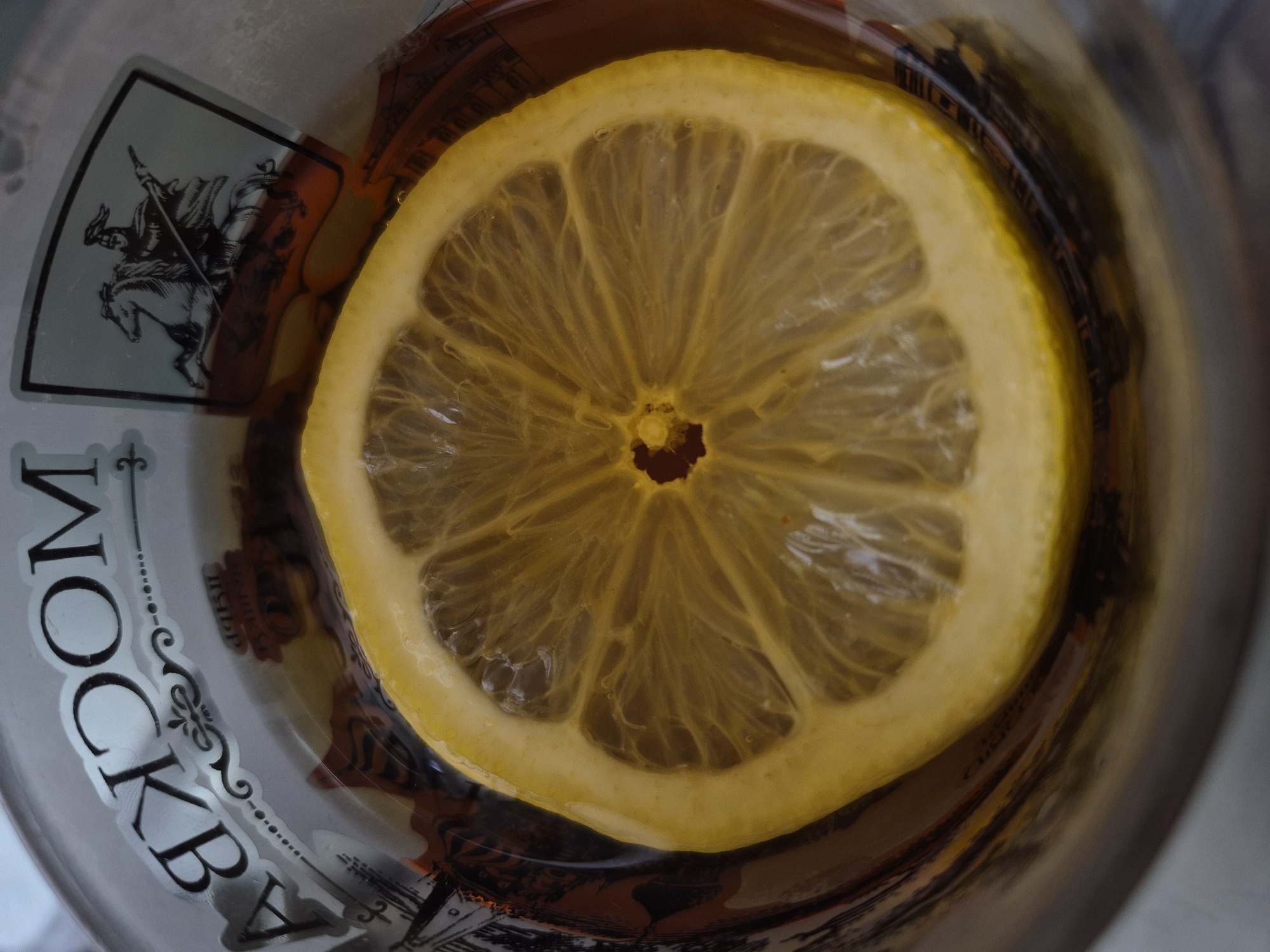 A close-up, top-down shot captures a slice of lemon submerged in a liquid within a glass. The lemon slice, positioned centrally, exhibits a vibrant yellow hue. The liquid in the glass is dark. The glass is adorned with a label that reads "mockby" and displays an image of a knight on horseback. The environment is not clearly identifiable.