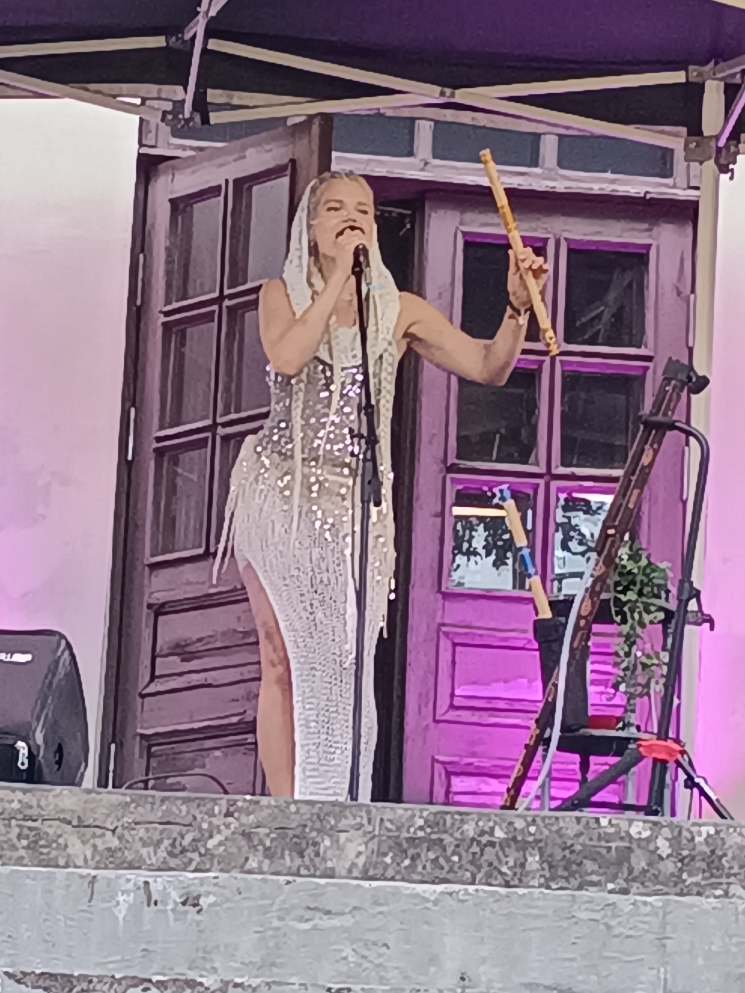 A woman wearing a sequined, white, high-slit dress and a white headscarf stands on a stone stage in front of a purple-hued wooden door with four panels. She is holding a microphone in her right hand and a wooden stick in her left. A black speaker is to her left, and a black stand with decorative plants and a wooden instrument is to her right. The scene is illuminated by purple stage lights. This appears to be an outdoor performance, possibly at the Aizpute Castle Courtyard in Aizpute, Latvia.