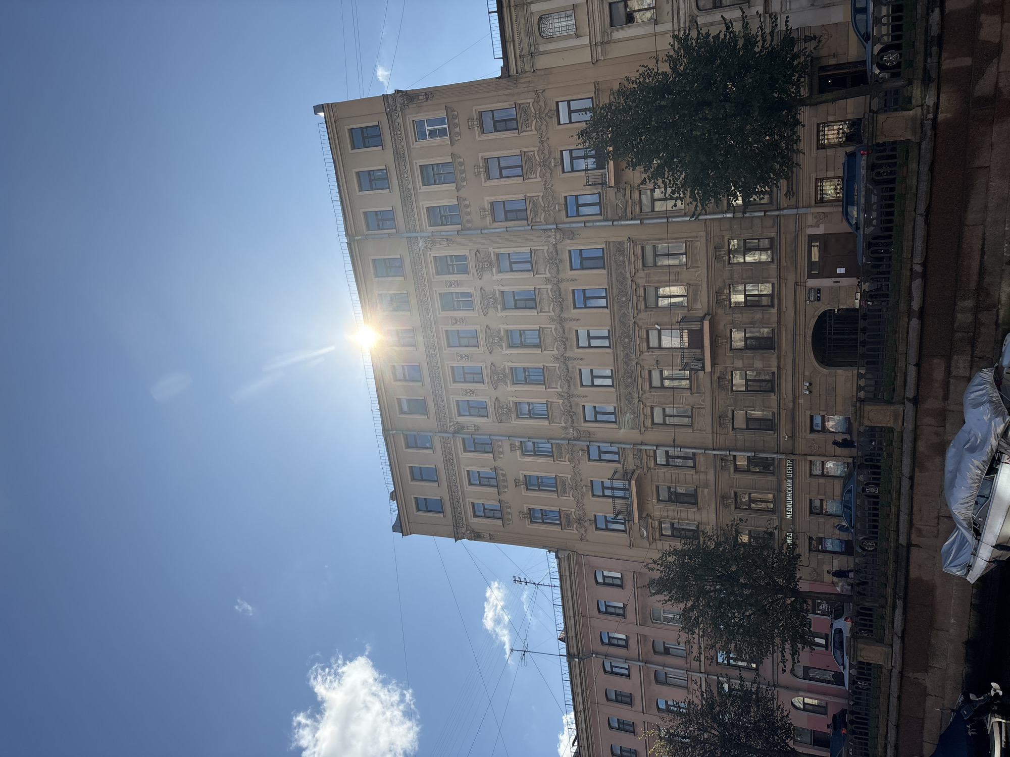 The image shows a sunlit view of buildings lining a canal in Saint Petersburg, Russia. The architecture is typical of the city, with ornate facades and multiple stories. The sun is bright, casting lens flare and illuminating the blue sky with scattered clouds. Several cars are parked along the street in front of the buildings, and a few people are walking on the sidewalk. A metal railing separates the sidewalk from the canal, where boats are docked. The overall scene conveys a sense of a pleasant day in a historic urban setting. Specifically, the building with the sign "МЕДИЦИНСКИЙ ЦЕНТР" indicates it is likely a medical center.