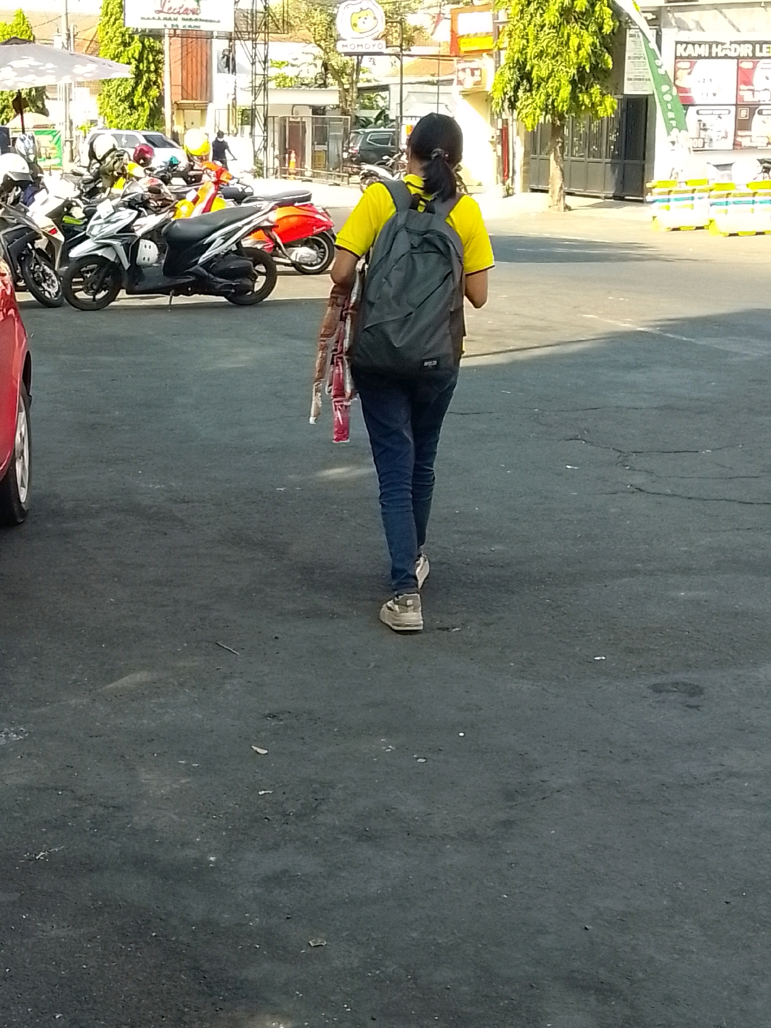 A person wearing a yellow shirt and blue jeans walks away from the camera, carrying a grey backpack and a string of red and white items. The scene takes place on a paved outdoor area, likely a parking lot, with several motorcycles parked in the background. Buildings and trees are visible beyond the parking lot. The presence of a red car on the left side of the frame suggests it is a public area. The image appears to be taken in Jember, Indonesia, as indicated by the context provided.