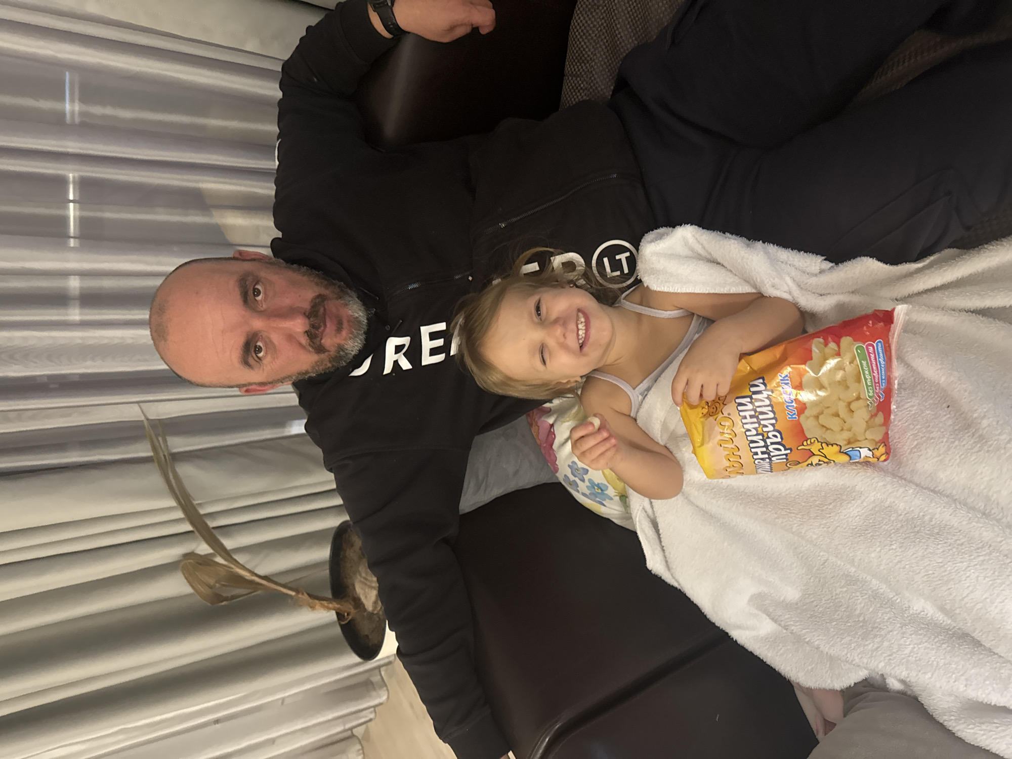 A man and a young girl are lying on a dark-colored sofa, possibly inside a residential building. The man has a shaved head and a beard and is wearing a black hooded sweatshirt. The girl is wrapped in a white towel and is holding a package of snacks. A plant is visible on the left side of the image. A curtain is behind them. The location is unknown and there is no explicit information to suggest the photo was taken in Varna, Bulgaria.