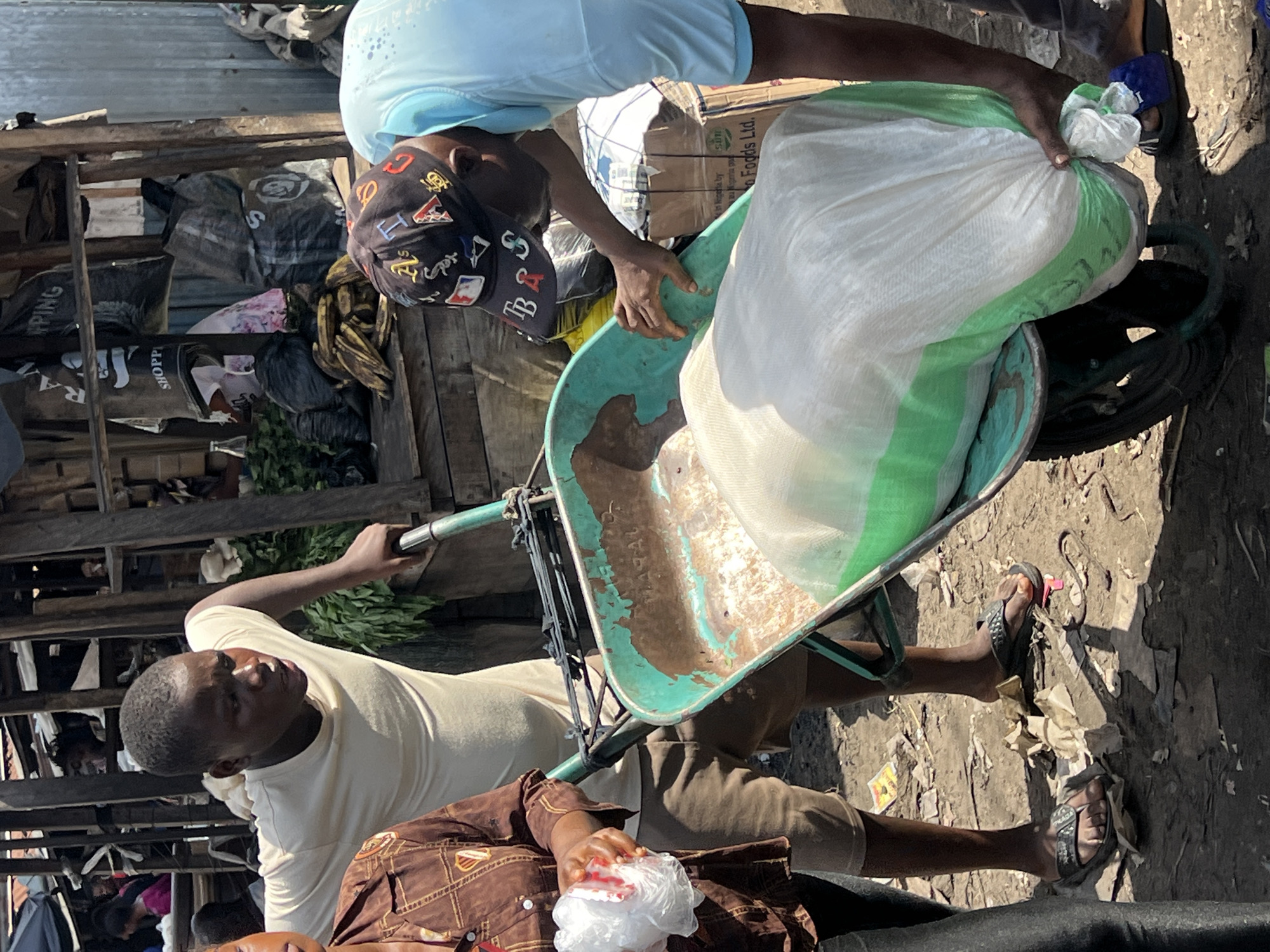 The image depicts a scene in Nkpor, Nigeria, showing two men loading a large white bag with a green stripe into a weathered, teal-colored wheelbarrow. The wheelbarrow appears to have some mud or dirt inside. One man, wearing a light blue shirt and a baseball cap with multiple emblems, is leaning over the wheelbarrow, presumably placing the bag inside. The other man, wearing a cream-colored shirt and tan shorts, is holding the handles of the wheelbarrow, looking forward. A third individual is partly visible, wearing a brown shirt, seated or positioned in front of the wheelbarrow. The ground surrounding the wheelbarrow is littered with various items, including trash and debris. In the background, there is a wooden structure with bags and other objects visible. The scene suggests an outdoor market or working environment.