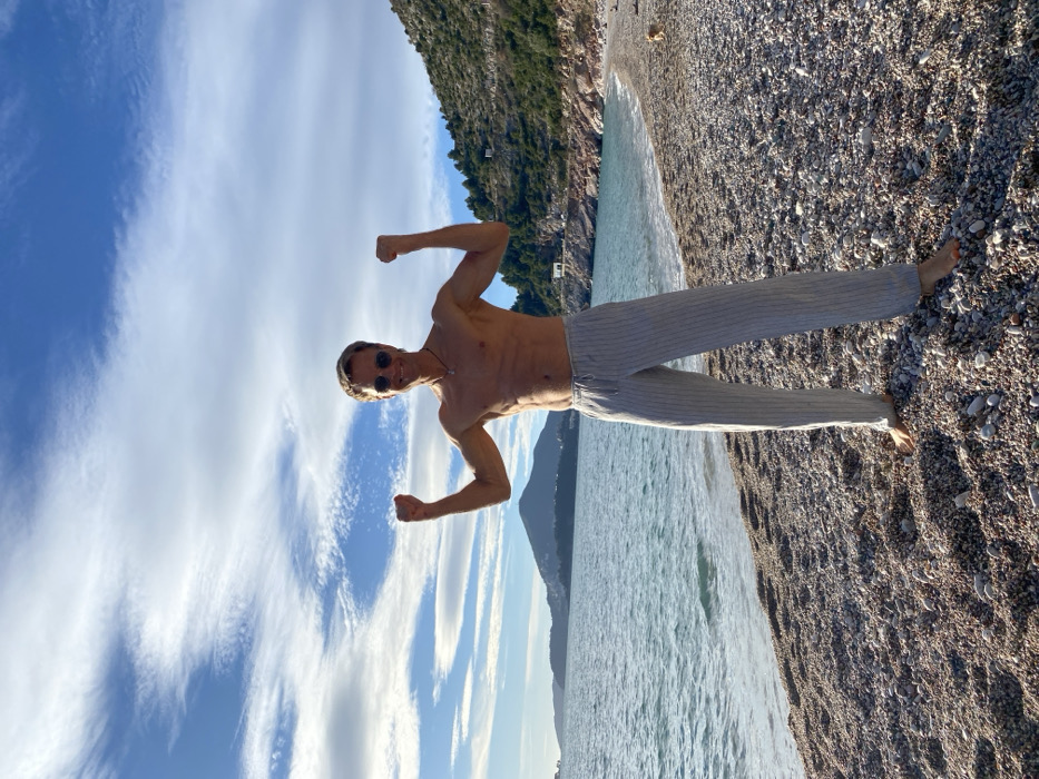 A man is captured in a full-body shot on a sunny pebble beach next to a body of water, likely the sea. The image is oriented vertically with the man standing upright, facing slightly towards the camera with a confident, smiling expression. He appears to be in his 30s or 40s, with a lean, muscular physique, and is shirtless, showcasing his torso. He wears light-colored, possibly off-white or light grey, striped full-length trousers. His bare feet are visible on the pebbles. He is wearing dark sunglasses and a thin necklace around his neck. His arms are bent at the elbows, with fists clenched, in a classic flexing pose, highlighting his biceps.
Behind him, to his right, is a wide expanse of clear, light blue-green water with gentle ripples. The water meets the shore, which is a wide pebble beach extending into the foreground. In the mid-ground and background, dark green and rocky mountains or large hills rise from the water's edge on the left side of the frame. A small, light-colored building is visible on the slope of one of these hills. The sky above is a vibrant blue, filled with numerous delicate, wispy white clouds (likely cirrus or altocumulus), indicating clear weather. The overall impression is one of bright sunshine, suggesting either late morning or early afternoon. The setting appears to be a picturesque coastal landscape, likely a holiday or leisure spot. No text is visible in the image.