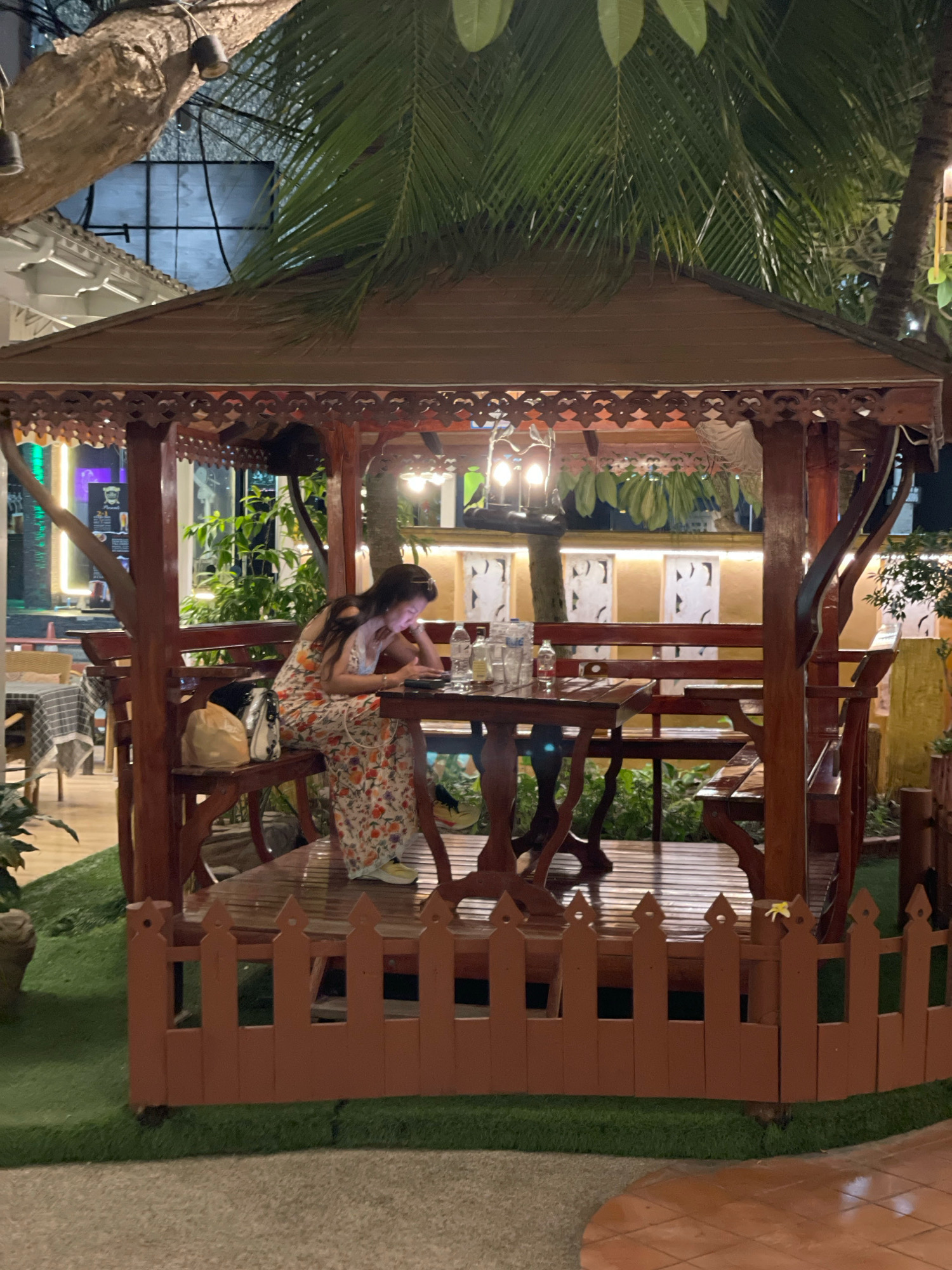 The image depicts an outdoor seating area at night, likely a restaurant or cafe in Pattaya City, Thailand. A woman is seated at a wooden table inside a wooden gazebo, looking down at her phone. She is wearing a floral dress. On the table are several water bottles and possibly a plate. A bag is placed on the bench next to her. The gazebo is surrounded by artificial grass and a low, red wooden fence. In the background, trees and palm fronds are visible, along with some outdoor lighting and building structures, suggesting an urban setting.