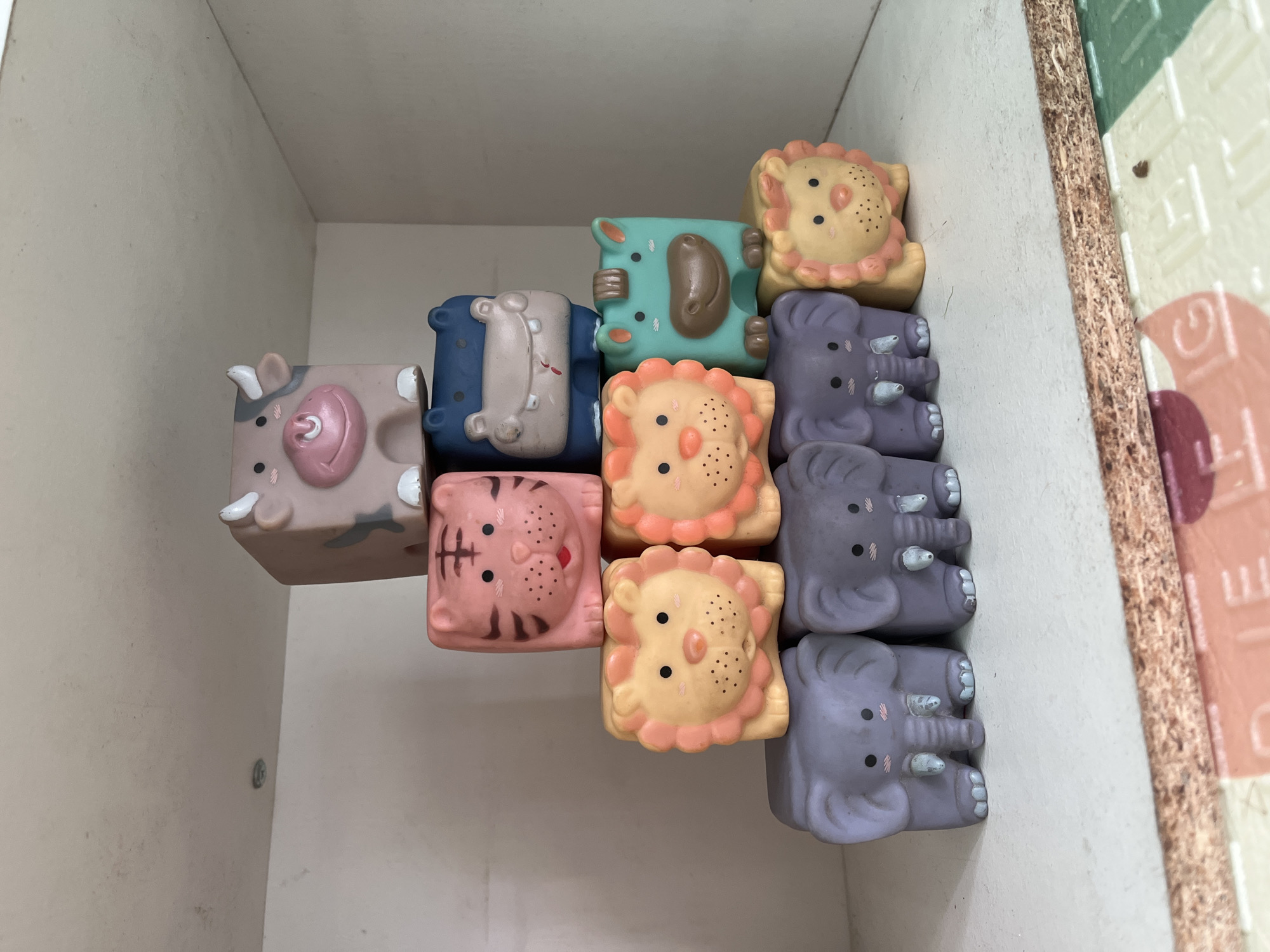 The image shows a pyramid of nine toy animals stacked on a shelf. The toys are made of soft plastic and depict cartoonish versions of a cow, tiger, hippo, horse, and elephants. The cow is at the top, followed by the tiger and hippo, then the horse and two lions, and finally a row of three elephants at the bottom. The toys are brightly colored and appear to be in good condition. The shelf is white and appears to be made of wood. There is some dust visible on the shelf. The background is a white wall. The lighting in the image is soft and even. The scene appears to be indoors, in a room that might be a child's bedroom or playroom.
The location of the image is not Kuta, Indonesia. There is no information in the image to suggest this location. The scene is a domestic setting, likely a shelf within a home.