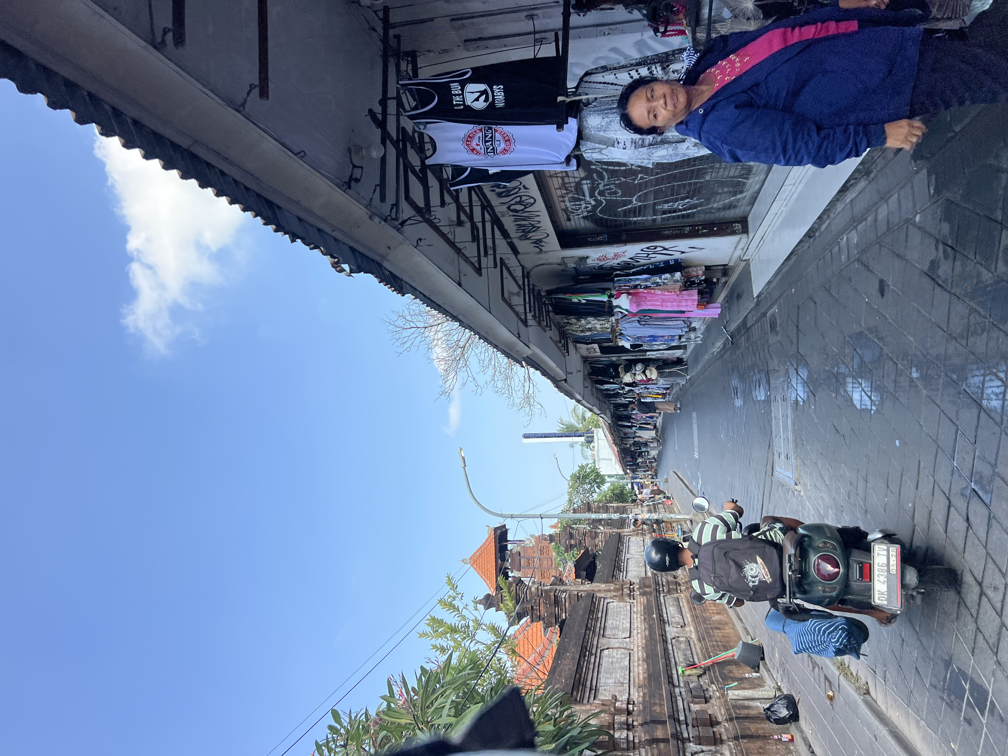 A street scene in Kuta, Indonesia, with a narrow cobblestone road flanked by shops on the right and a brick wall with traditional architecture on the left. A person wearing a black helmet and a striped shirt is riding a scooter away from the camera, with a backpack and a blue striped bag on the back. The shops display a variety of clothing items hanging from racks. A woman in a blue jacket and pink shirt stands in front of one of the shops, looking towards the right. The sky is blue with scattered clouds.