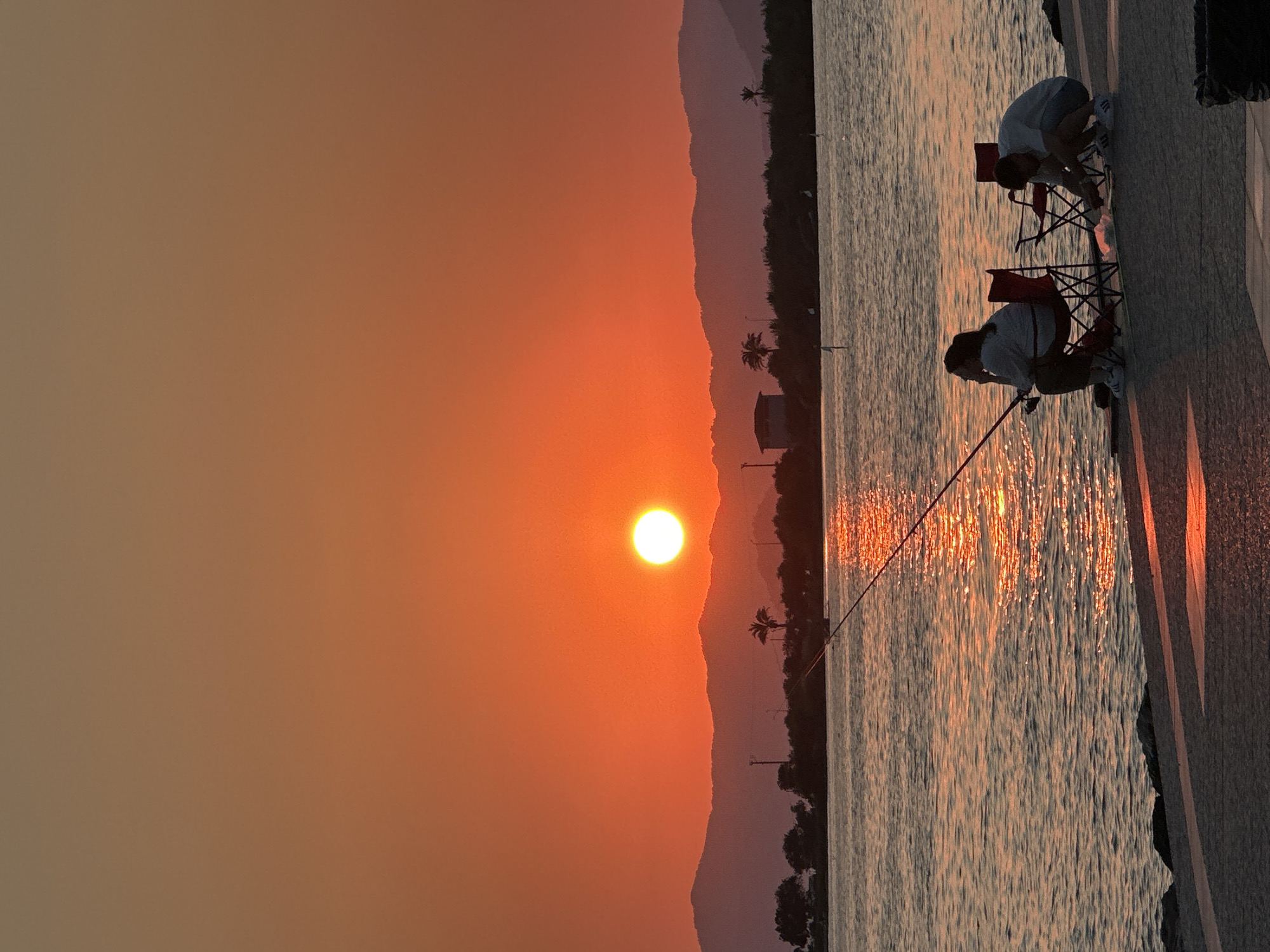 The image depicts a sunset over the water in Fethiye, Turkey. The sun, a bright orange orb, is low on the horizon, casting a warm glow across the sky and reflecting on the water. In the foreground, two people are sitting on chairs by the water, fishing. They are silhouetted against the sunset, making their features indistinguishable. The water is calm, with gentle ripples. In the background, there are hills and trees, also silhouetted against the sky. The overall impression is one of peace and tranquility.