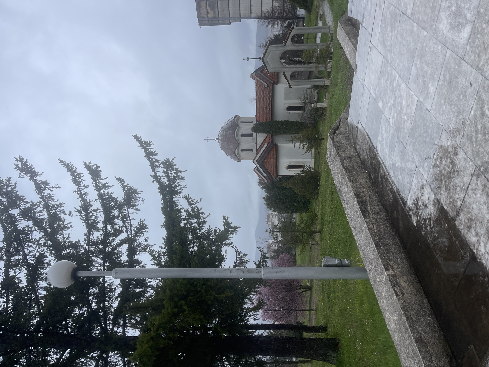The image captures a view in Veliko Tarnovo, Bulgaria, featuring the Holy Forty Martyrs Church. The church, white with a red tile roof and a dome topped with a cross, sits amidst a grassy area with trees. A small bell tower stands adjacent to the church. To the left, a tall coniferous tree and a streetlight are visible. In the foreground, the edge of a stone structure, possibly steps or a monument, is prominent. The sky is overcast, contributing to a muted color palette. A taller building is partially visible in the background.