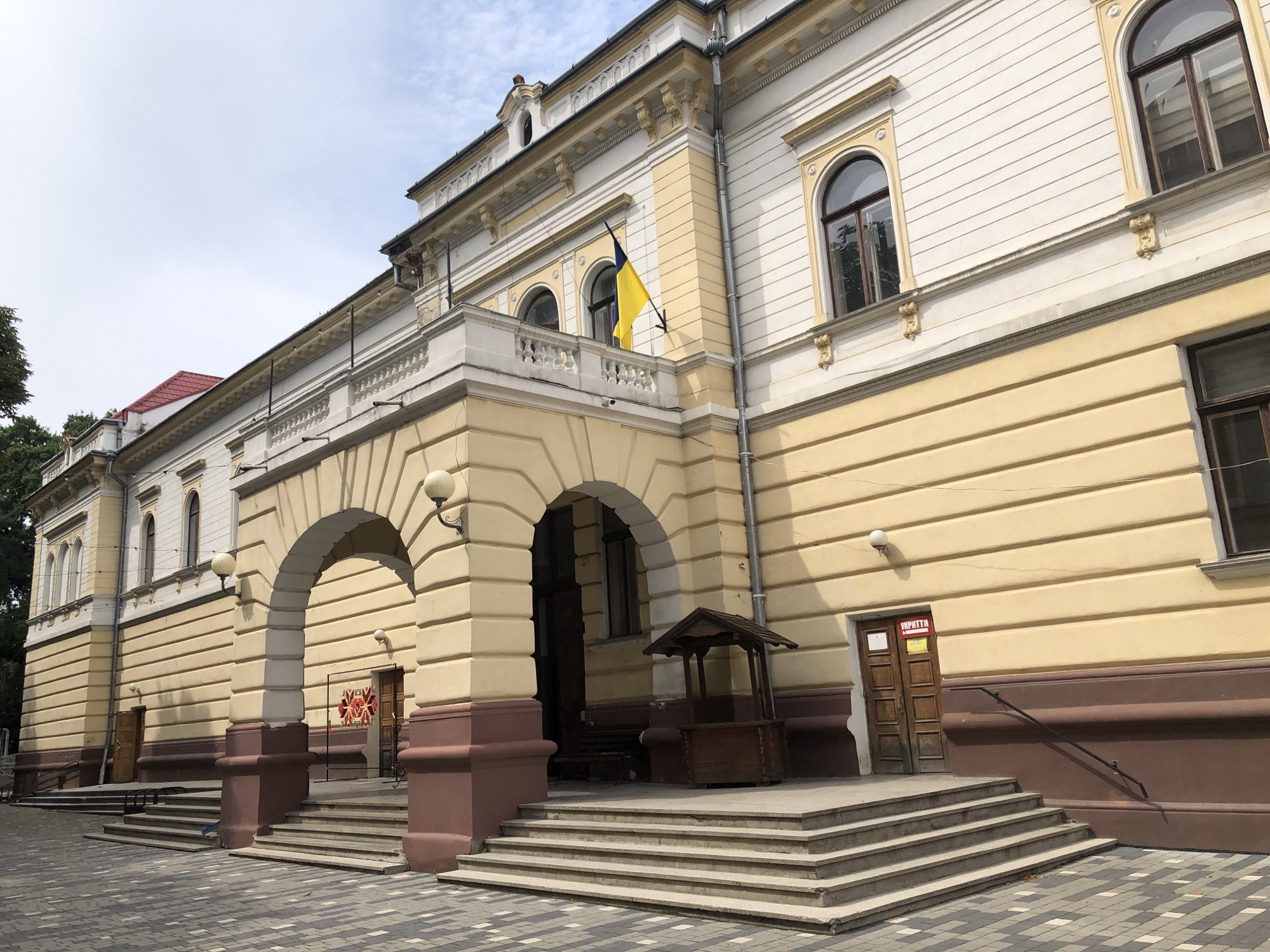 This is a full shot of a historical building located in Kolomyia, Ukraine. The building is multi-story and features a light yellow facade with white and dark brown architectural details. The building's design incorporates arched windows with dark frames and decorative molding. A Ukrainian flag is prominently displayed on a balcony on the second floor. The foreground shows a paved area with a patterned stone design, leading up to a wide staircase. Several wooden doors are visible, and a small wooden kiosk with a pitched roof stands near the entrance. The sky is overcast with a few clouds.