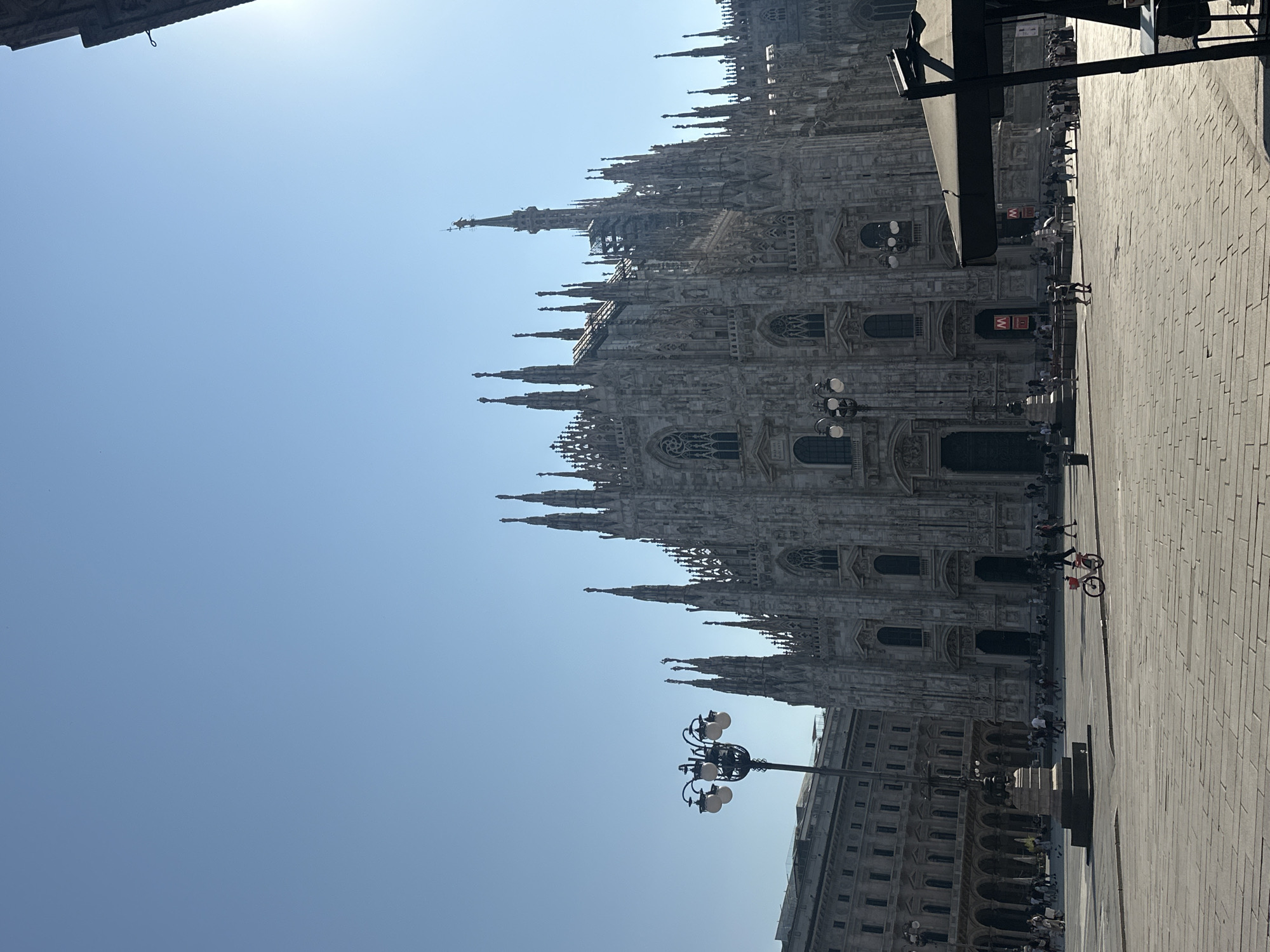 The image depicts a wide shot of the Piazza del Duomo in Milan, Italy, on a clear, sunny day. The magnificent Milan Cathedral, a Gothic architectural masterpiece, dominates the background, its intricate spires and facade bathed in sunlight. In the foreground, the square is paved with stone tiles, and scattered individuals are seen walking or standing. A vintage-style lamppost with multiple globes stands on the left side of the frame. To the left of the cathedral, a grand building with arched windows and colonnades is partially visible. On the right, a dark-colored umbrella-shaded table is partially in view. A red bicycle is parked in the middle of the square, and a few more people are spread throughout the plaza. The sky is a clear, bright blue with a slight glare from the sun.