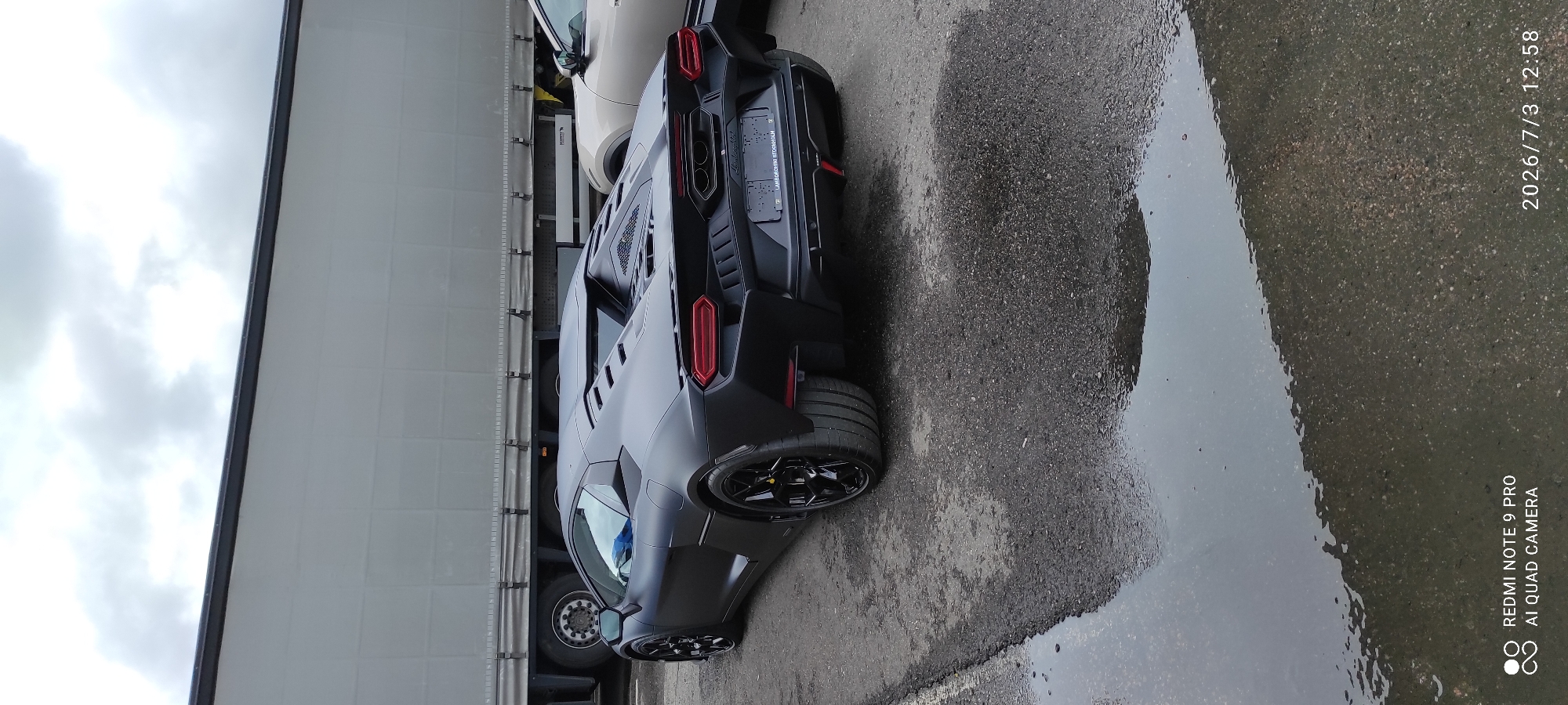 A matte black Lamborghini Veneno is parked on a wet asphalt surface. The car has a low profile, sharp body lines, and distinctive rear taillights. In the background, a large, grey, semi-trailer truck is visible. Another vehicle, a light-colored SUV, is parked to the right of the Lamborghini. The sky is overcast with clouds. There are no people visible in the image. The timestamp in the lower right corner of the image indicates the date and time are 2026/7/3 12:58. The text in the lower left corner states "REDMI NOTE 9 PRO AI QUAD CAMERA".
