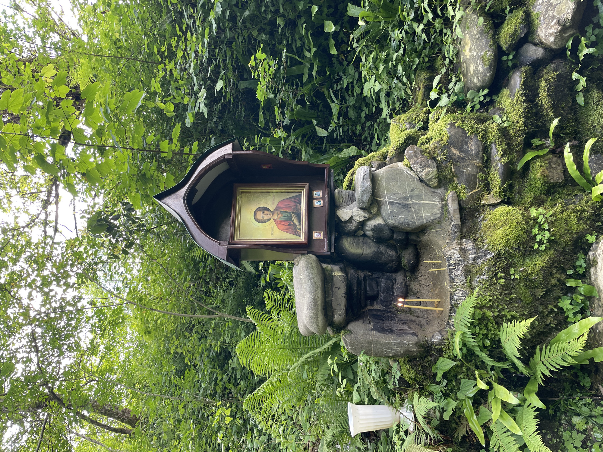 The image depicts a small, wooden shrine nestled amidst lush vegetation, likely in Solokh-Aul, Russia. The shrine contains a framed icon of a religious figure, possibly a saint, with a halo. Below the icon, two lit candles stand on a stone structure covered in moss. The surrounding environment features a variety of green plants, including ferns and mosses, suggesting a damp, wooded area. The shrine is partially obscured by trees, giving the scene a secluded and natural feel.
