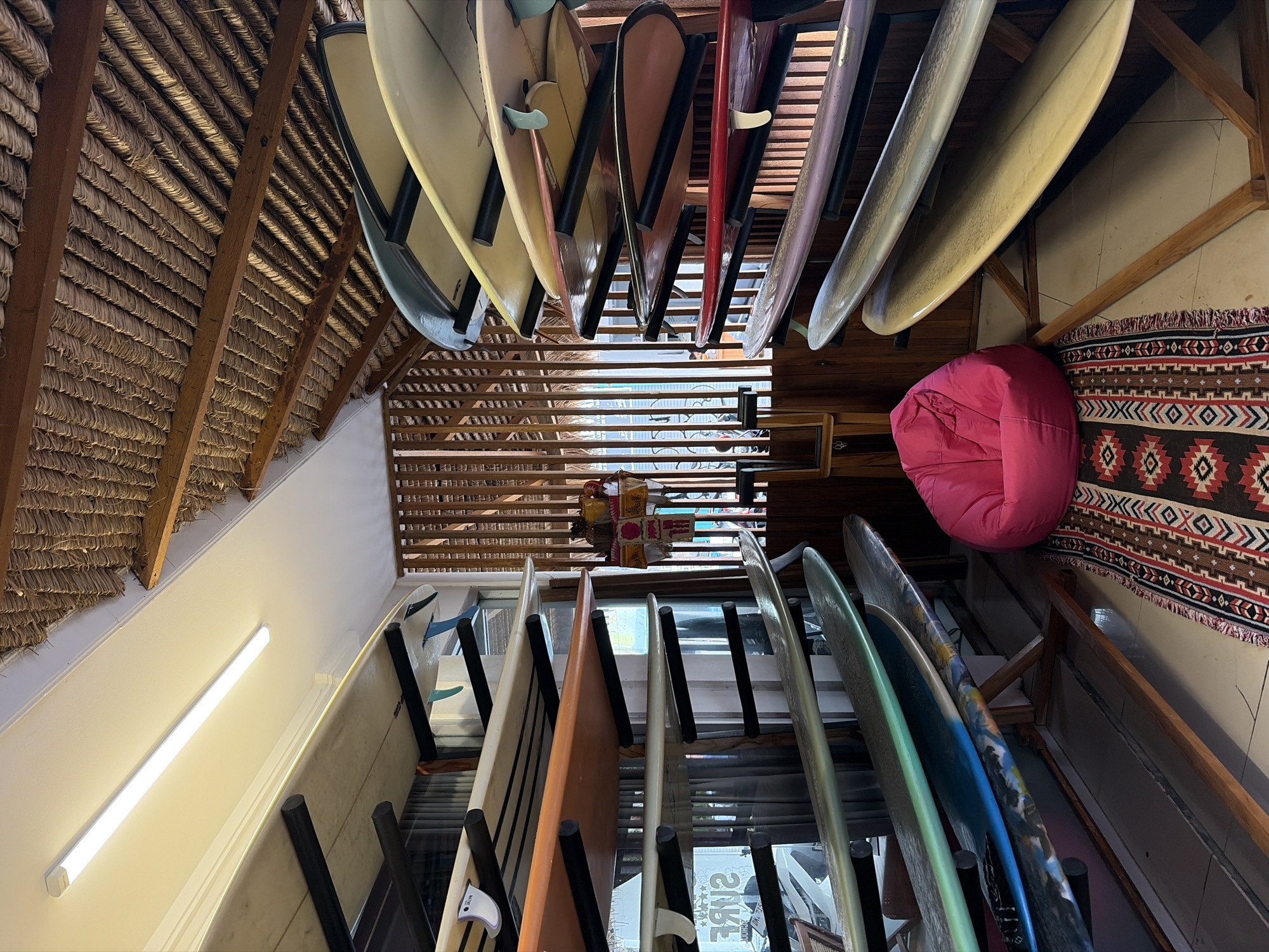 A brightly lit indoor surfboard storage facility in Kuta, Indonesia, features a collection of surfboards organized on wooden racks. The room has a thatched roof and wooden slat walls, creating a rustic ambiance. A pink beanbag chair sits on a patterned rug in the foreground, and a long fluorescent light fixture illuminates the scene.