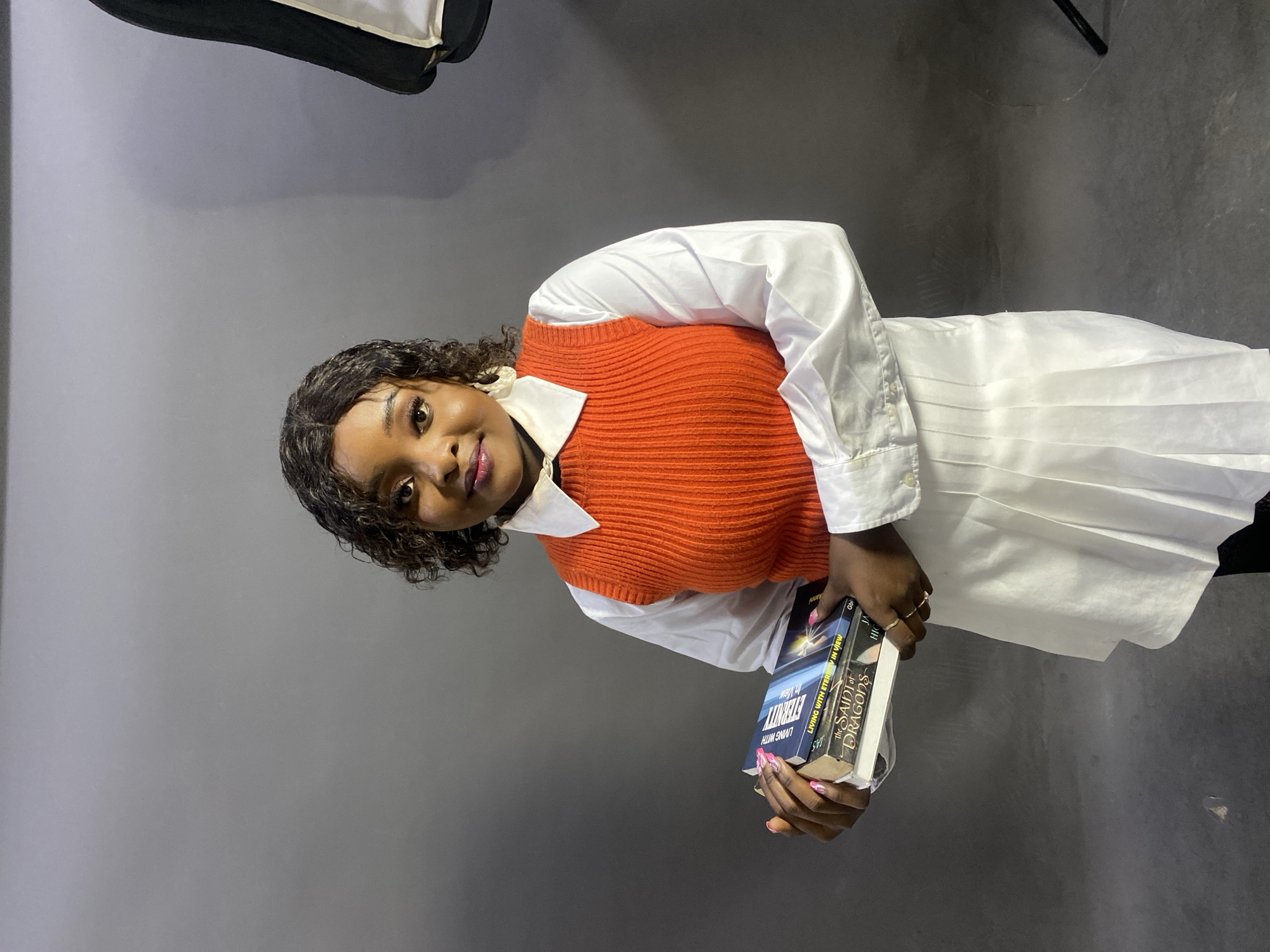 A young woman with dark hair and skin stands in a studio against a grey background. She wears a white button-down shirt with an orange knitted vest. She holds three books in her hands. From left to right, the book titles and authors are "LIVING WITH ETERNITY IN VIEW" by Gbi, "LIVING WITH ETERNITY IN VIEW" by Gbi, and "The SAINT of DRAGONS" by JA. A light box is visible in the upper left corner. The location is Jos, Nigeria.