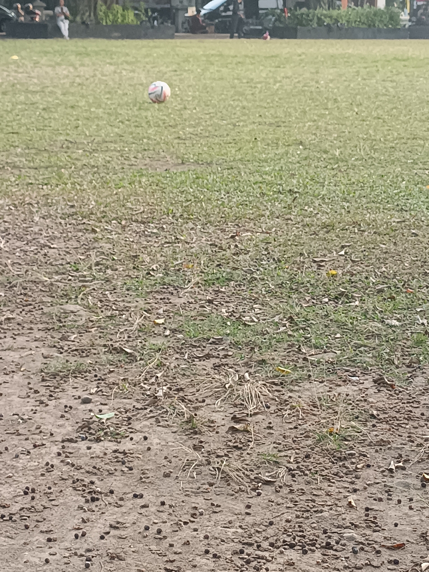 An outdoor, ground-level, eye-level shot shows a soccer ball in mid-air on a grassy field in Sleman, Indonesia. The ball, which is white with red, blue, and black accents, is positioned in the upper left quadrant of the image. The foreground of the image consists of dry, bare earth with scattered pebbles and dry grass. The background features a green, well-maintained grassy field with a few indistinct figures and structures in the distance, suggesting a park or sports facility. The lighting suggests an overcast or diffused daylight.