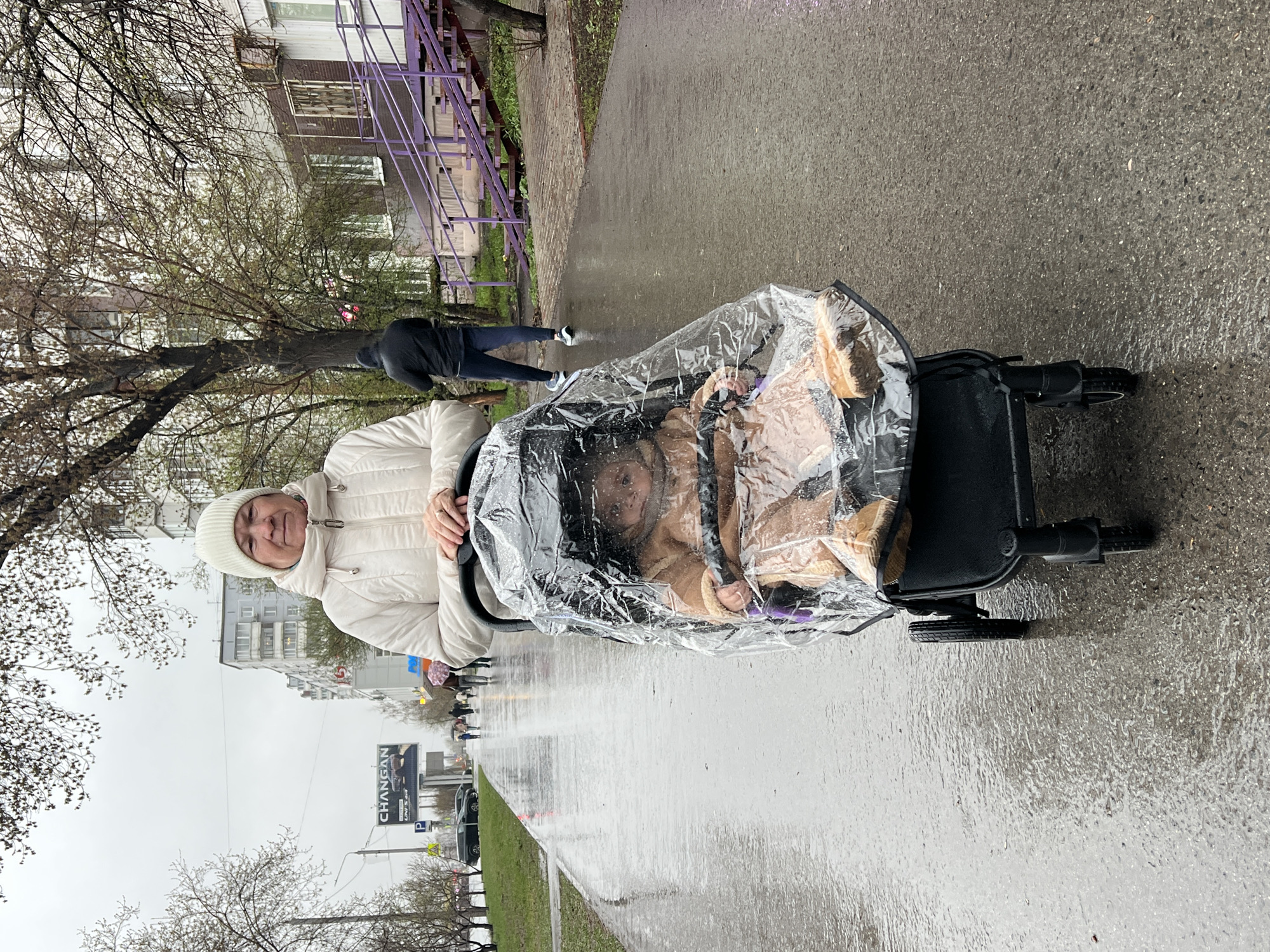 A woman in a white jacket and hat is pushing a stroller with a clear plastic rain cover on a wet sidewalk in Perm, Russia. The stroller appears to contain a child. The wet ground reflects the overcast sky and surrounding buildings. Trees with bare branches line the street, and a sign with the word "CHANGAN" is visible in the background. Other pedestrians and cars are present in the distance. A purple railing is seen alongside a building to the upper left.