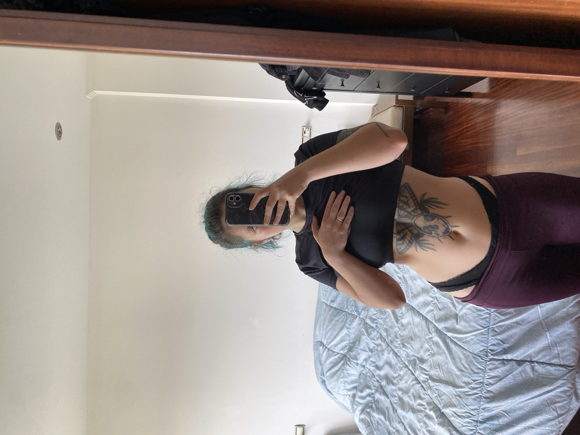 A woman is taking a selfie in front of a mirror. She is wearing a black crop top and purple leggings. She has a tattoo on her stomach. The mirror reflects a bedroom with a bed and a dresser. The location is unknown.
