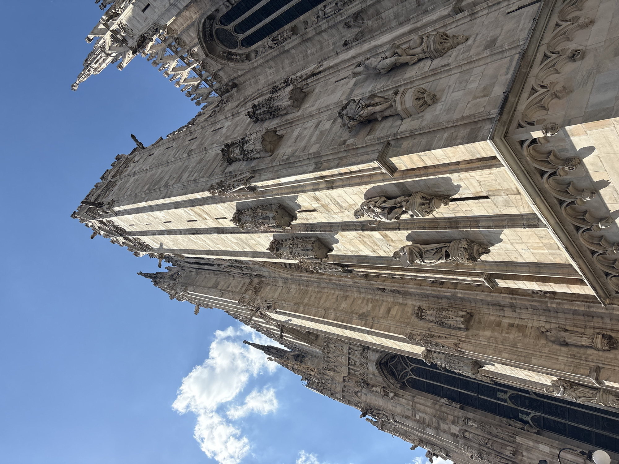 The image features a low-angle view of the Milan Cathedral (Duomo di Milano) in Milan, Italy. The architecture is characterized by white stone and elaborate Gothic details including spires and statues adorning the facade. The sky is blue with a few clouds. No people are visible in the frame.